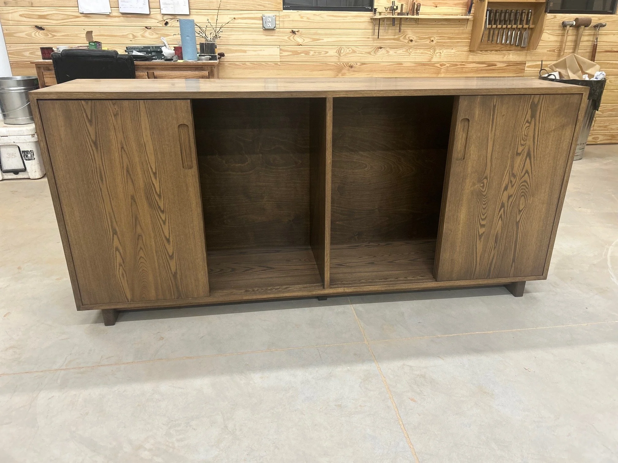 A wooden sideboard with sliding doors in a workshop. The sideboard has a natural finish and is empty inside, with a set of hardware tools on the wall above it.