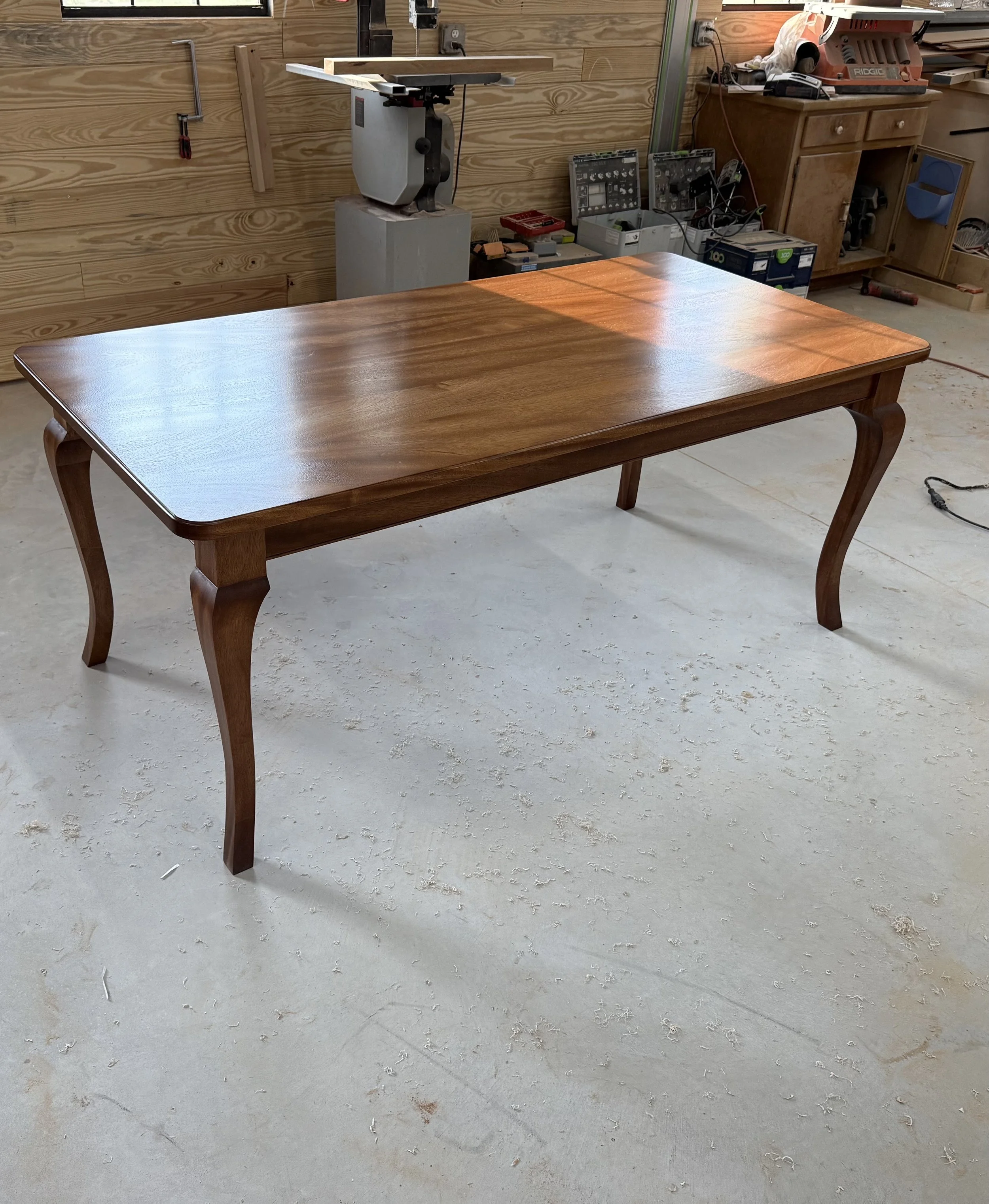 A polished wooden dining table with curved legs in a woodworking workshop.