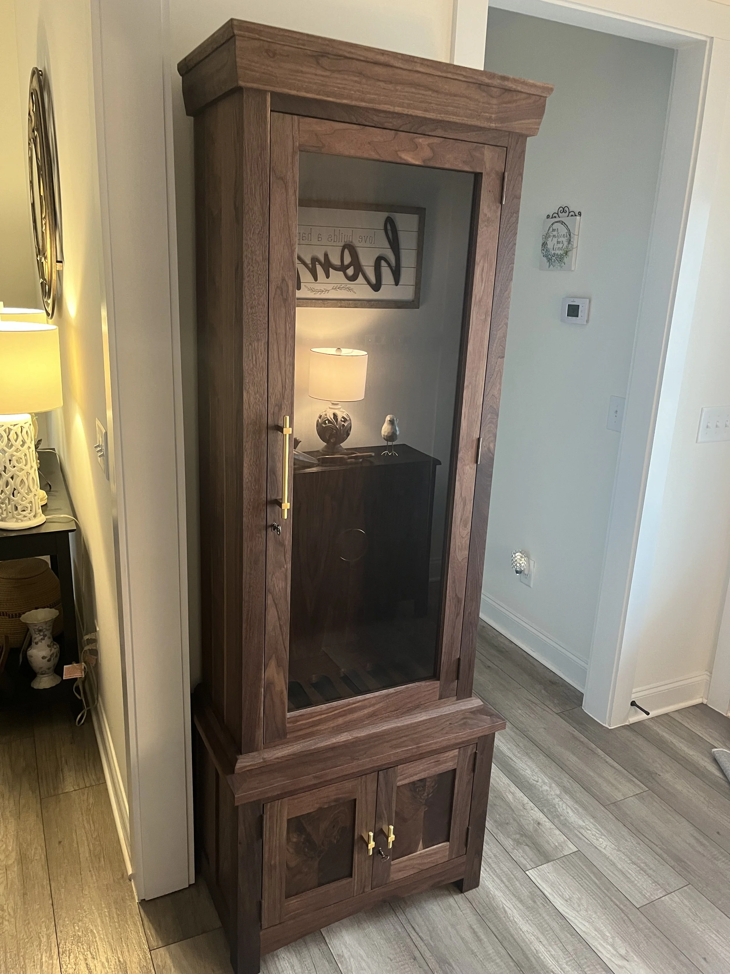 A wooden cabinet with glass door and lower storage, placed in a room corner, with decorative household items and a lamp reflected in the glass.