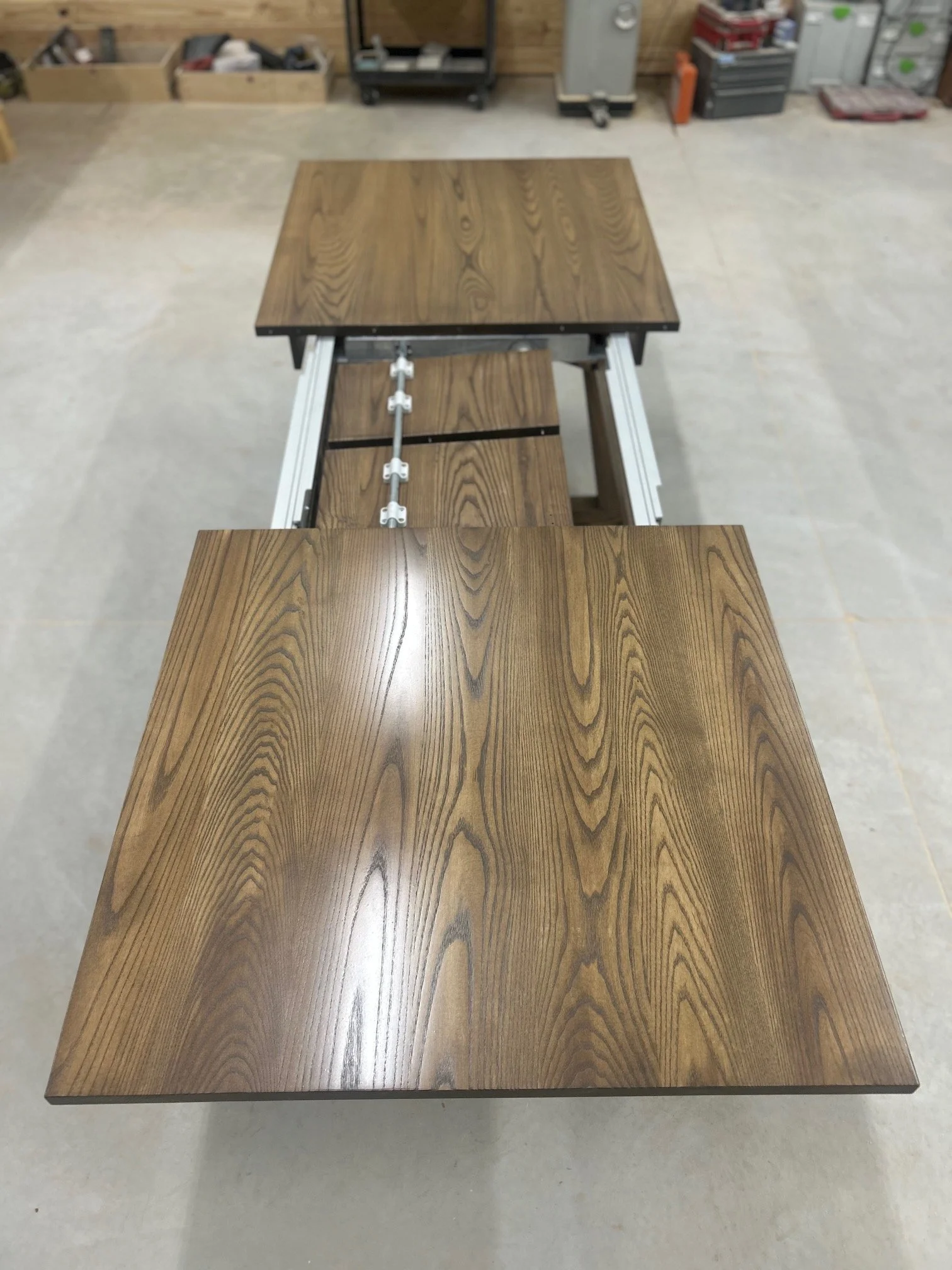 A woodworking workbench in a workshop with two wooden tabletops, featuring a natural wood grain pattern, attached to a metal frame with adjustable components.