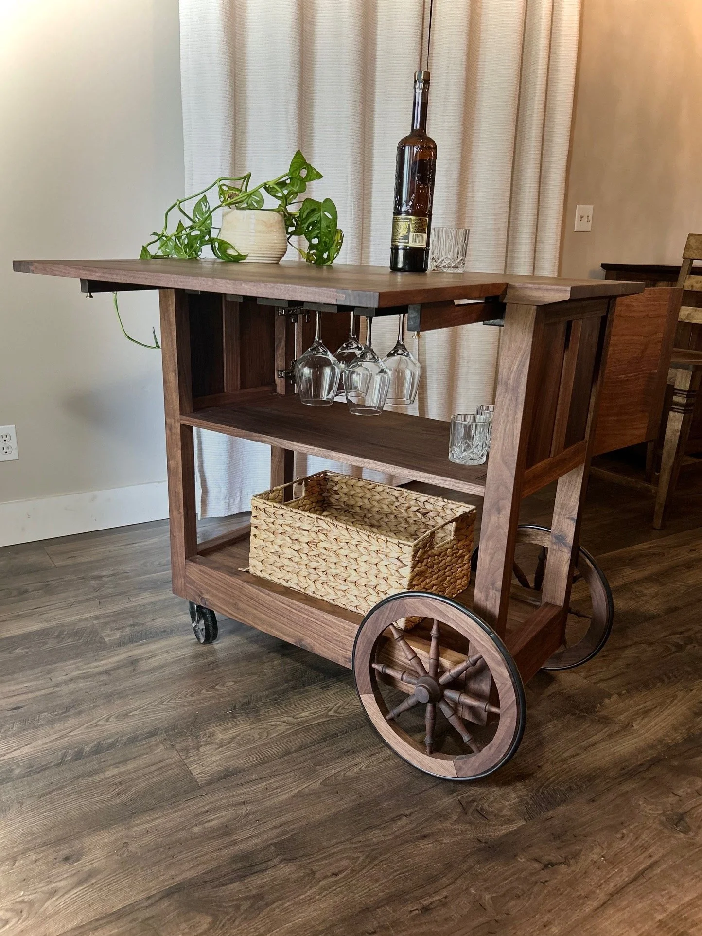 Wooden bar cart with wheels, holding a plant, bottles, glasses, and a woven basket, set on a wooden floor with curtains in the background.