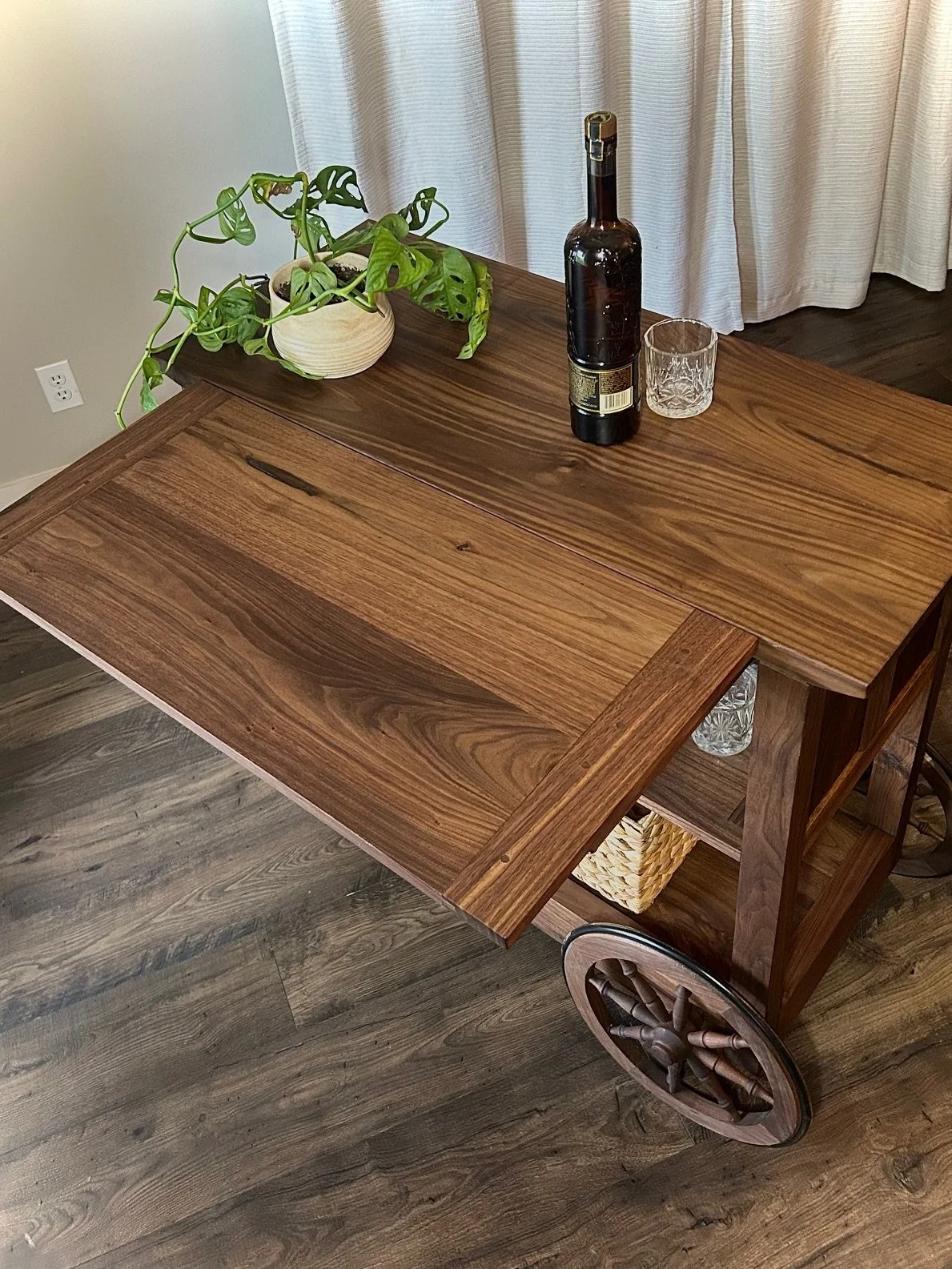 Wooden rolling cart with a large wooden tabletop, a potted plant with green leaves, a bottle with a wine label, a glass, and a basket underneath, in a room with hardwood floor and curtains.