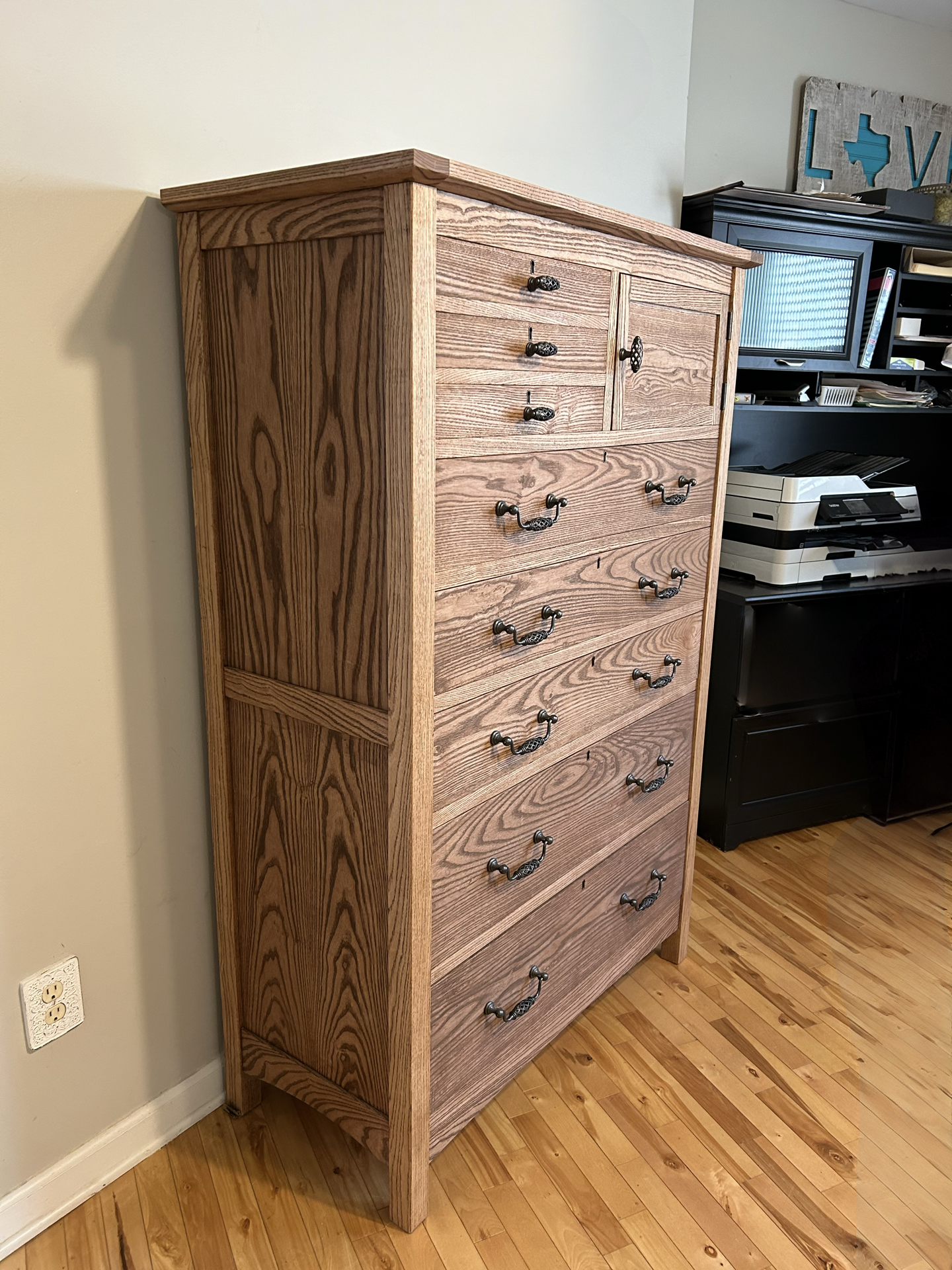 A wooden tall dresser with multiple drawers and vintage-style handles, situated in a room with wooden floor and a nearby black desk with office supplies.