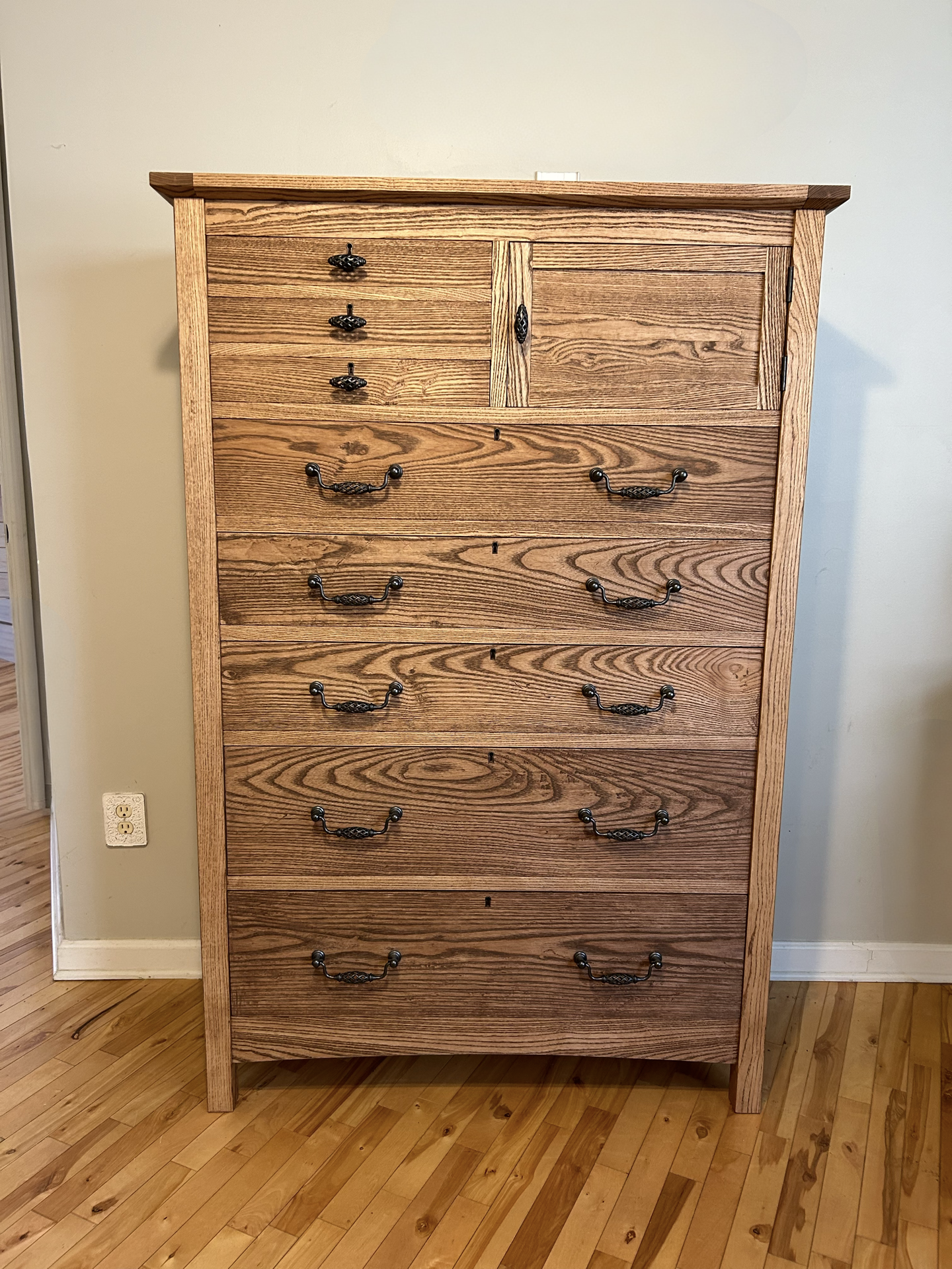 A tall wooden dresser with multiple drawers and black metal handles, placed against a beige wall, on a hardwood floor.