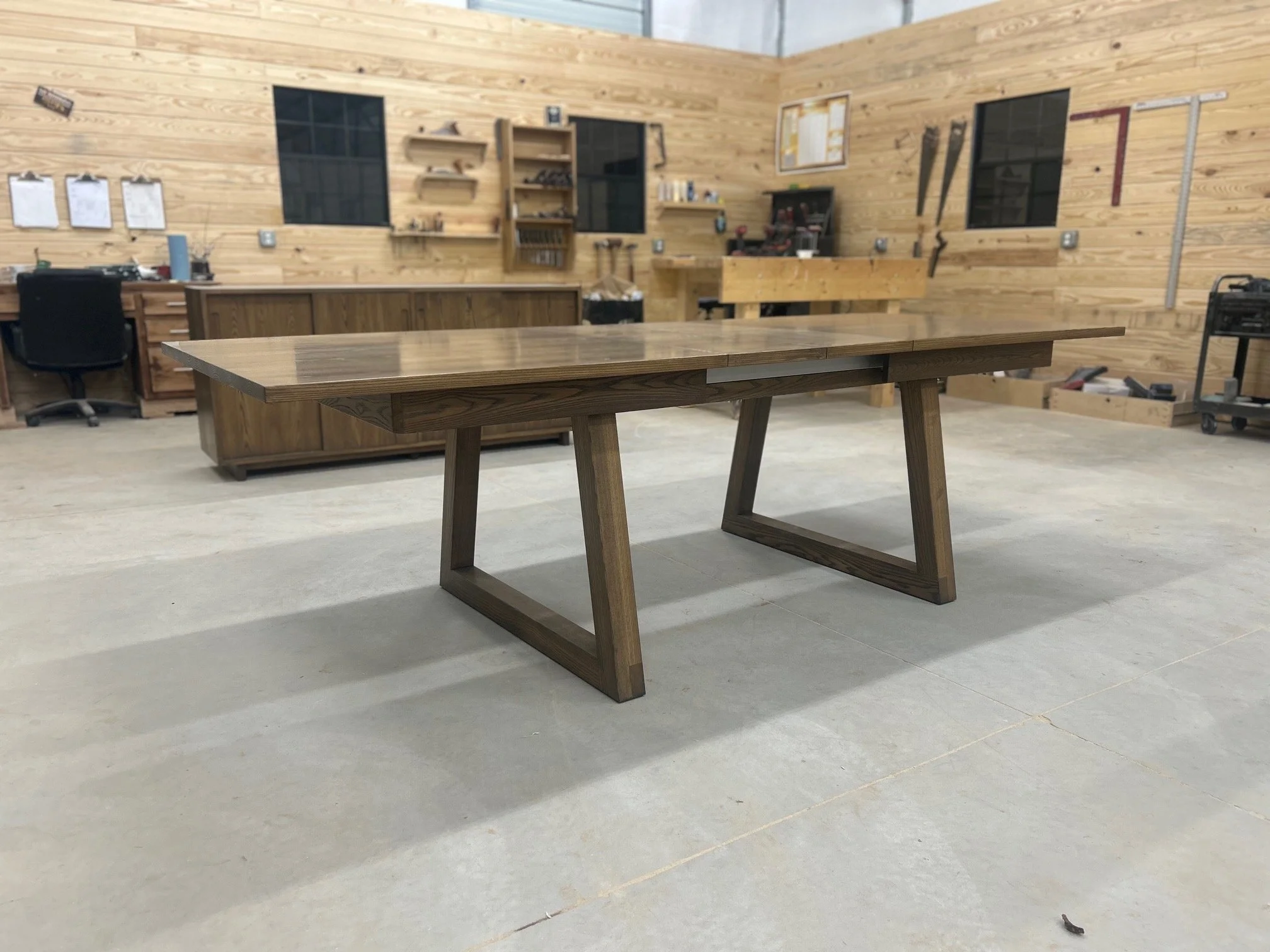A wooden table with angled legs in a woodworking shop with wooden walls, a workbench, tools, and storage shelves.