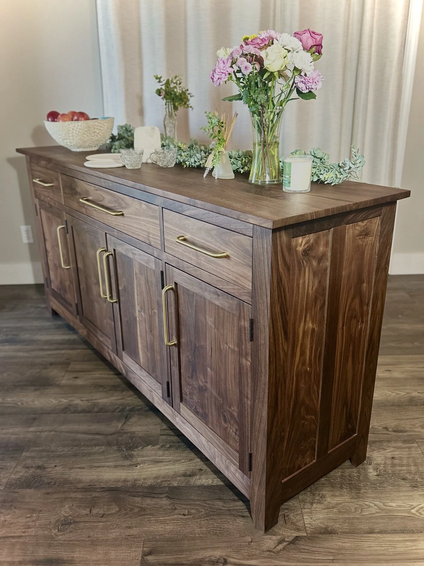 Wooden sideboard decorated with a large glass vase of pink and white flowers, a candle, and small decorative items, set against a beige curtain.