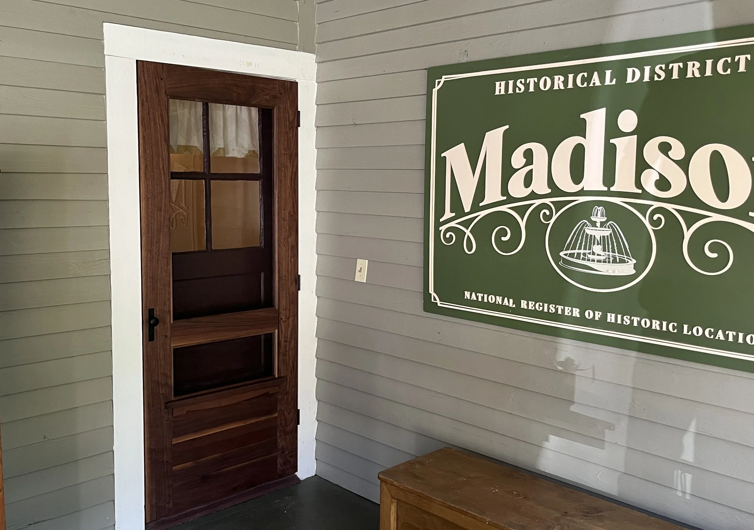 Wooden door next to a green sign that reads 'Madison, National Register of Historic Locations,' along with a fountain illustration, on a beige wall.