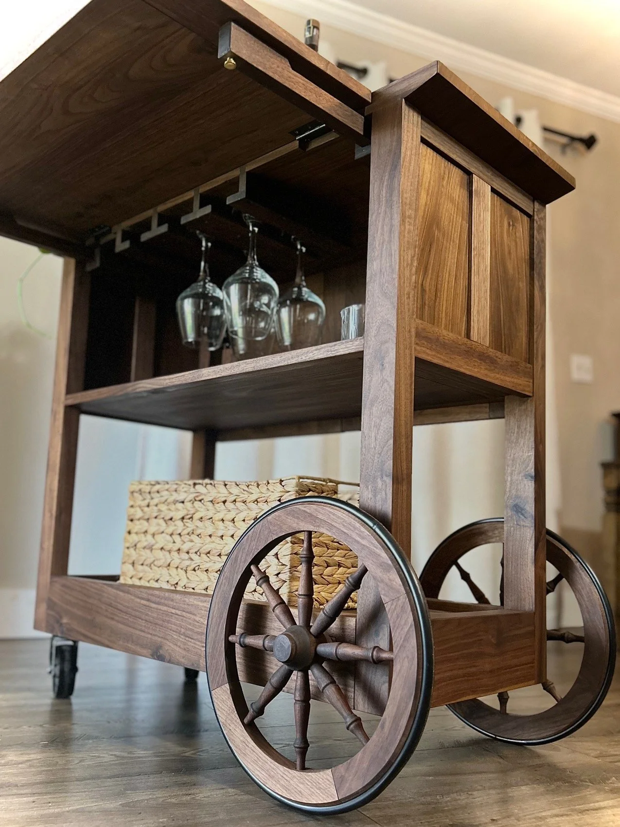 Wheeled wooden bar cart with wine glasses hanging upside down, basket on bottom shelf, wooden wheels, and a wood finish.