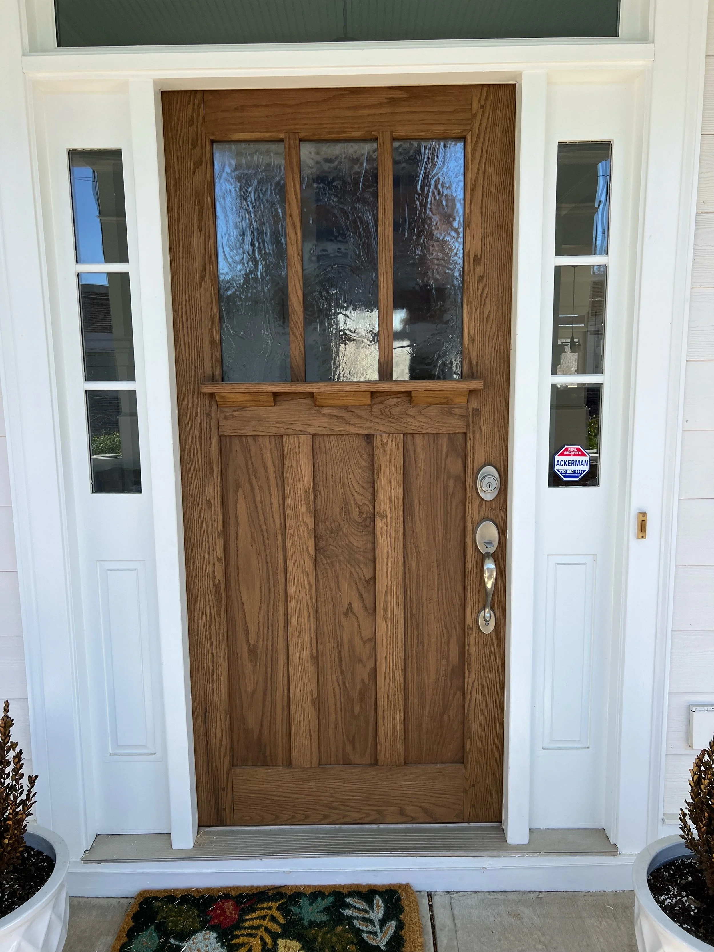 Front door made of wood with glass panels at the top, white framing, security sticker on the right side, and two potted plants on either side, with a decorative doormat in front.
