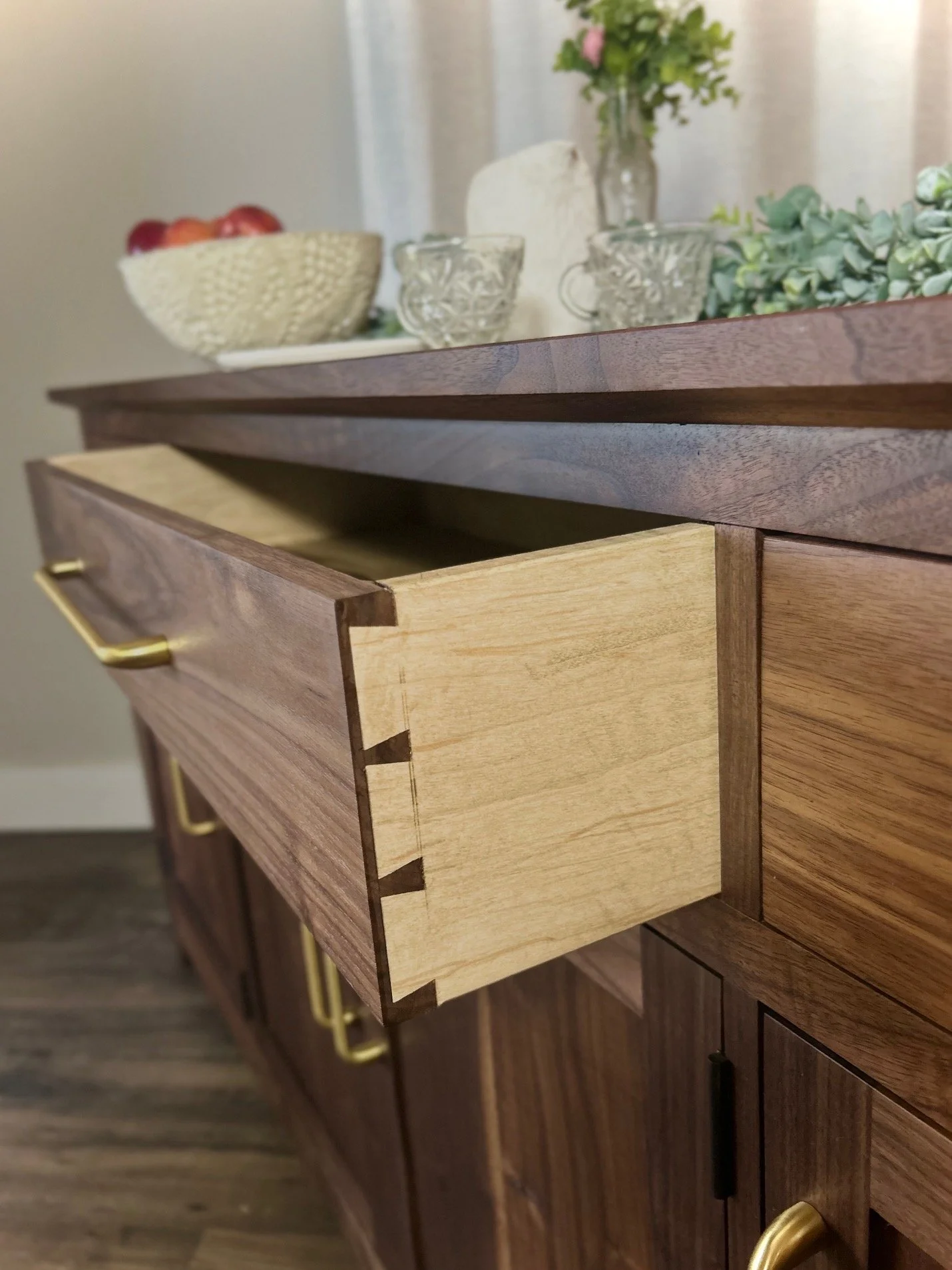 Close-up of a wooden dresser with a partially opened drawer, showing a contrasting light wood interior and dovetail joints. The top of the dresser has decorative glassware, a bowl of fruit, and flowers in a vase.