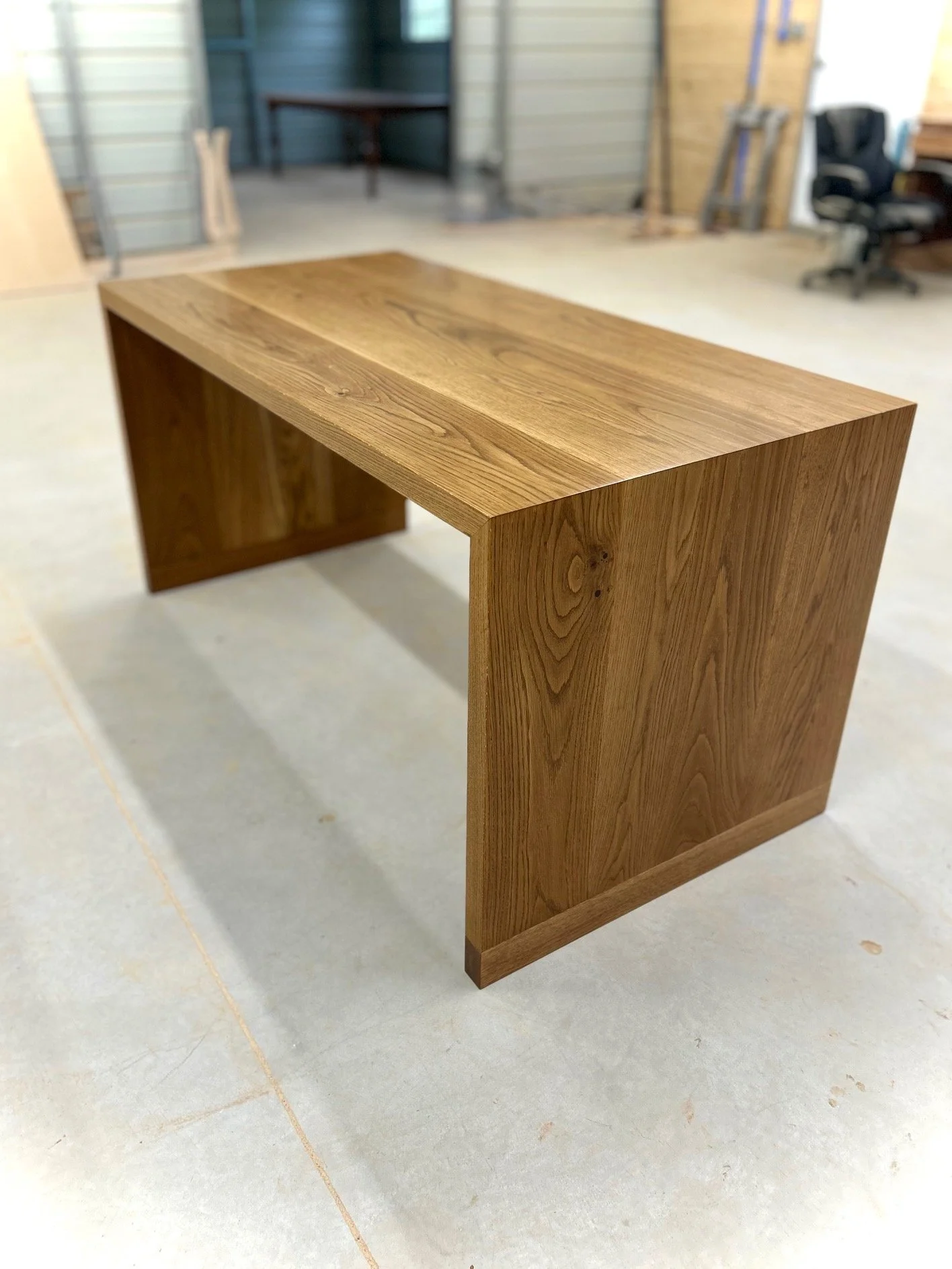 A wooden desk or table with a smooth, polished surface and sturdy legs, placed on a light-colored concrete floor in what appears to be a workshop or garage.