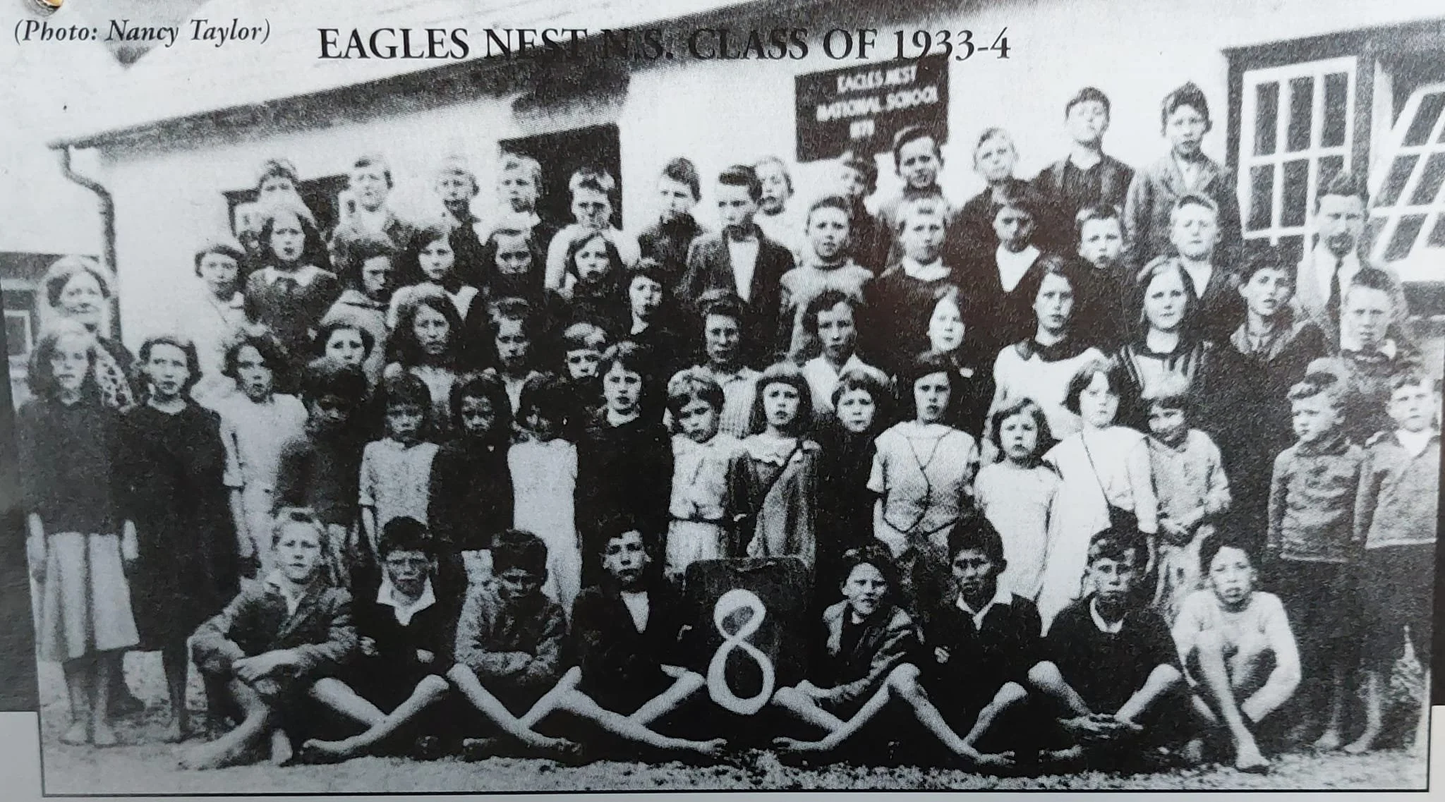 A black and white group photo of students and teachers from the 1933-1934 EAGLES NEST PRIMARY SCHOOL class, taken outdoors in front of the school building, with children standing and sitting, some seated on the ground and others on chairs, in front of a sign featuring a large number 8.