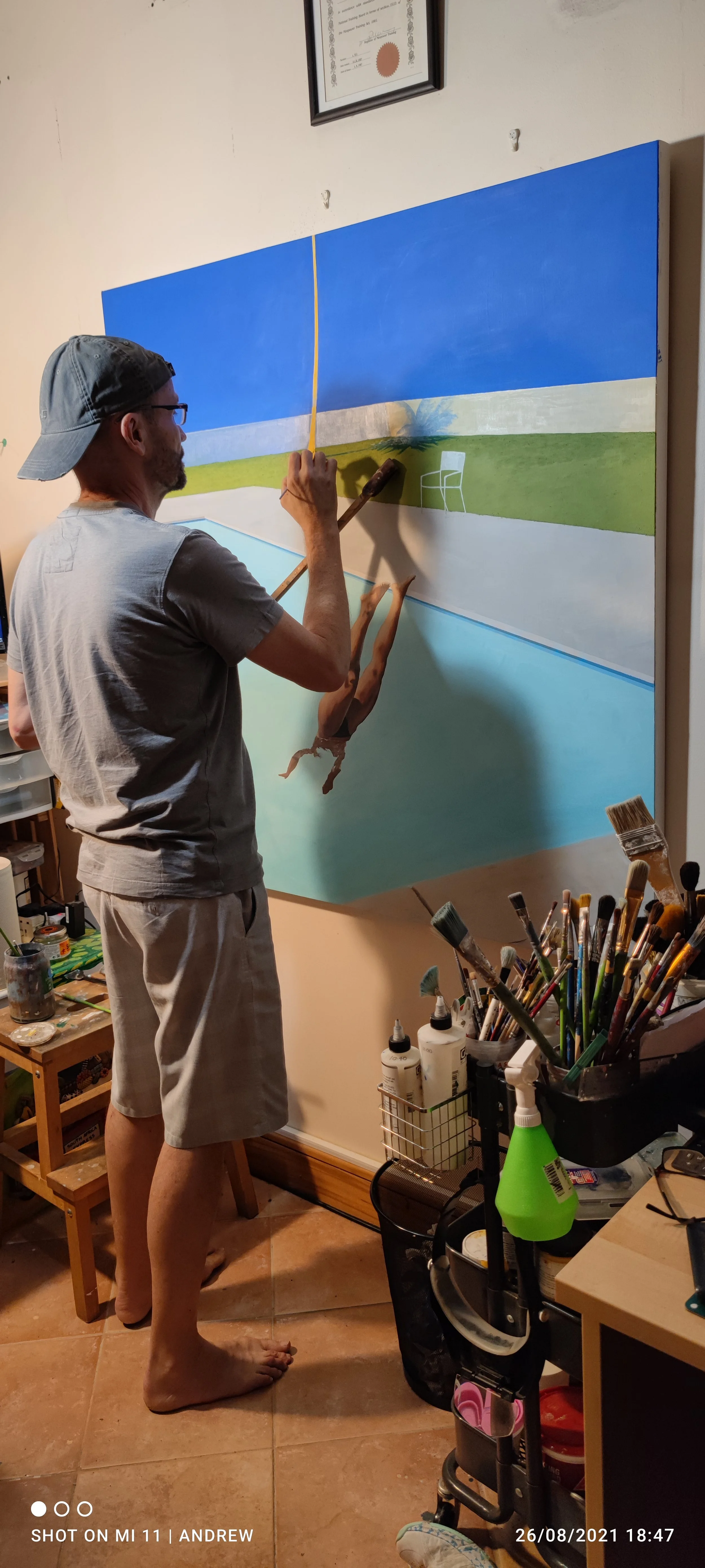 A man in a gray shirt, shorts, and a gray cap painting a landscape scene on a large canvas, with a blue sky, white clouds, green grass, and a chair visible in the background. The man is barefoot and working with a paintbrush and palette. Various painting supplies are on nearby tables.
