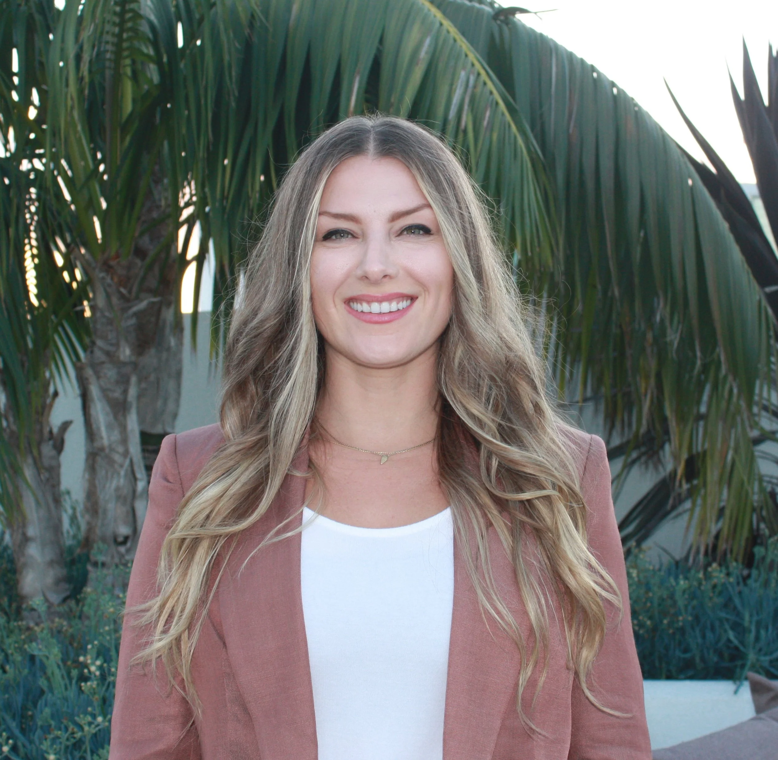 Chelsea wearing a brown blazer and white top standing outdoors with palm trees in the background.