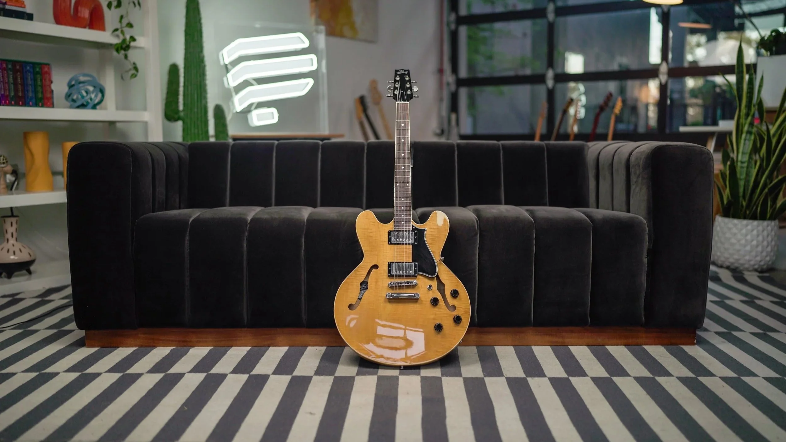A hollow-body electric guitar laying against a black sofa in a lounge. The Pickup Music logo is in the background and the guitar is waiting for you to pick it up and play.