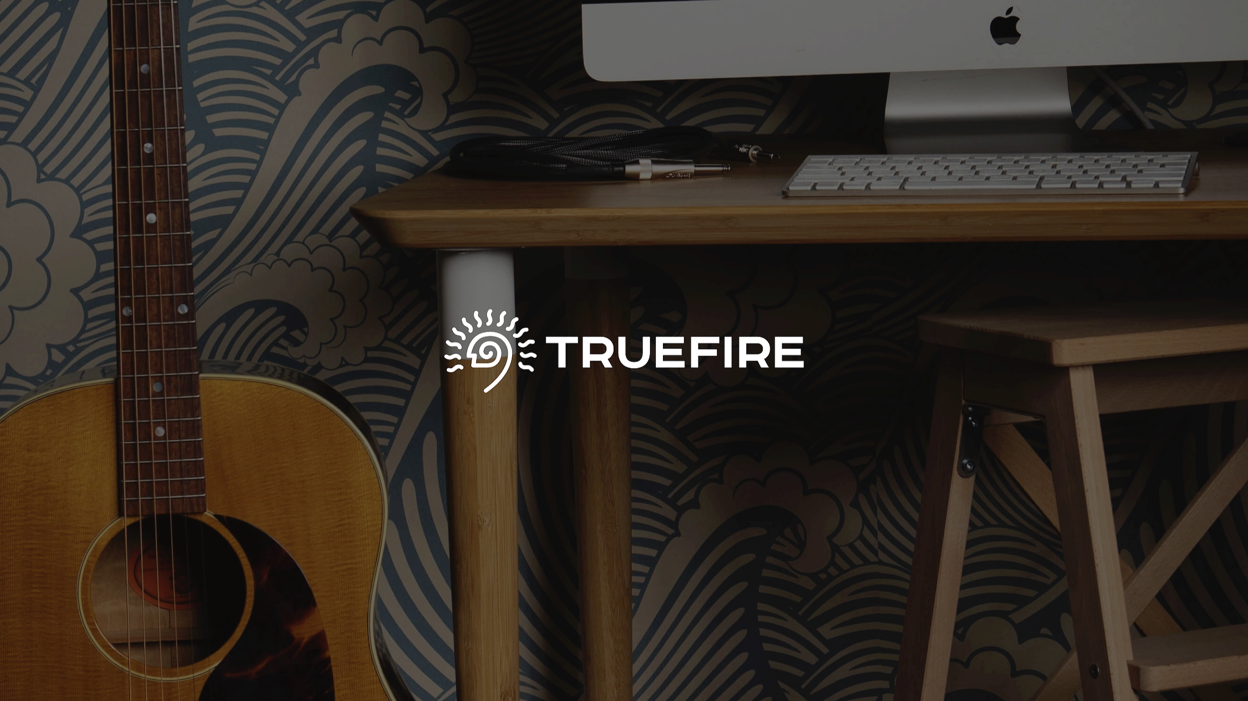 TrueFire logo overlayed on a background image of a music studio with an acoustic guitar and Apple Mac computer on a wooden desk with cables on top.