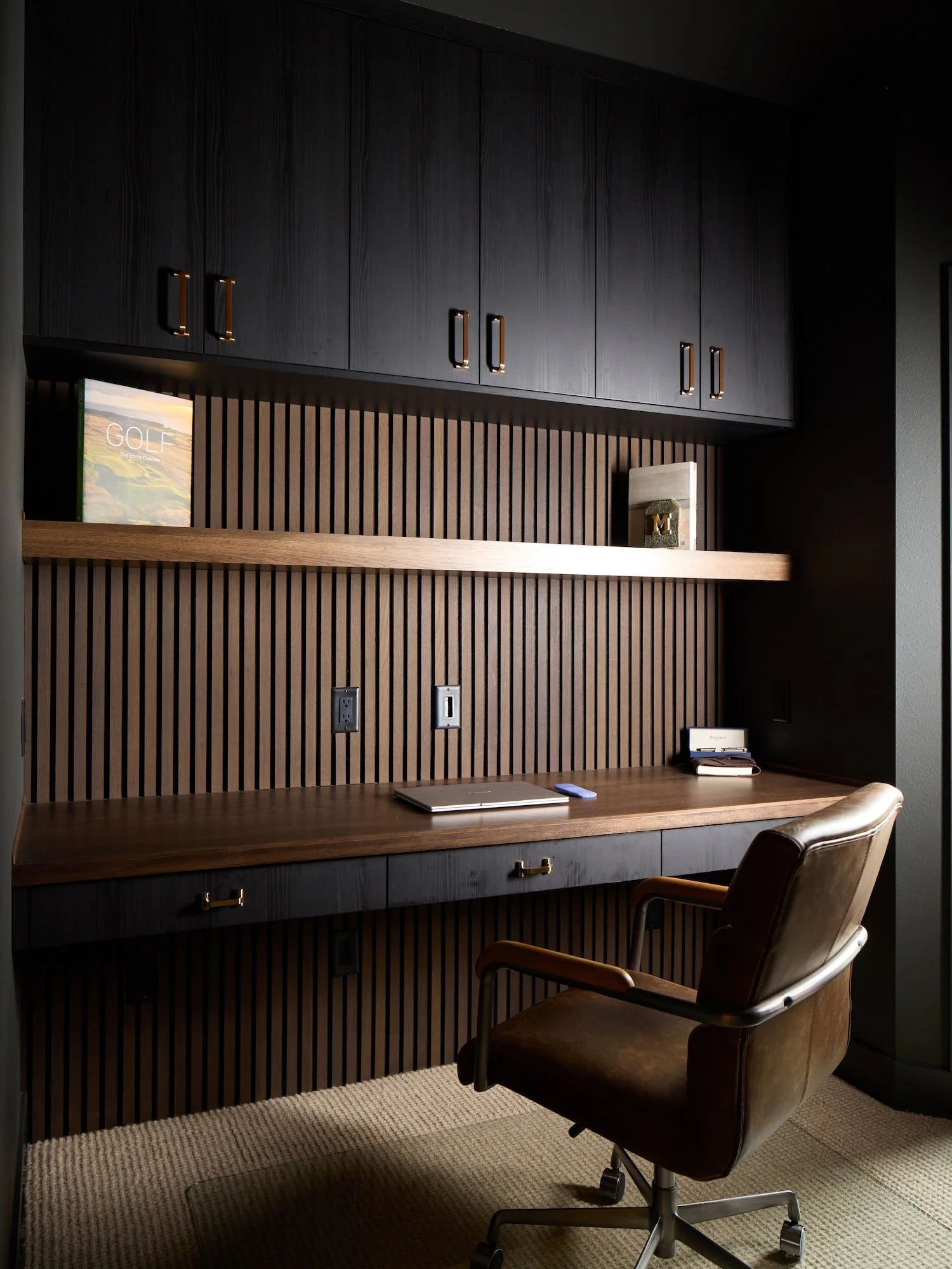 Modern office desk with a brown leather chair, a closed laptop, a phone, and a notepad. Dark wooden cabinets and a wooden shelf with decorative items are behind the desk, with electrical outlets along the wall.