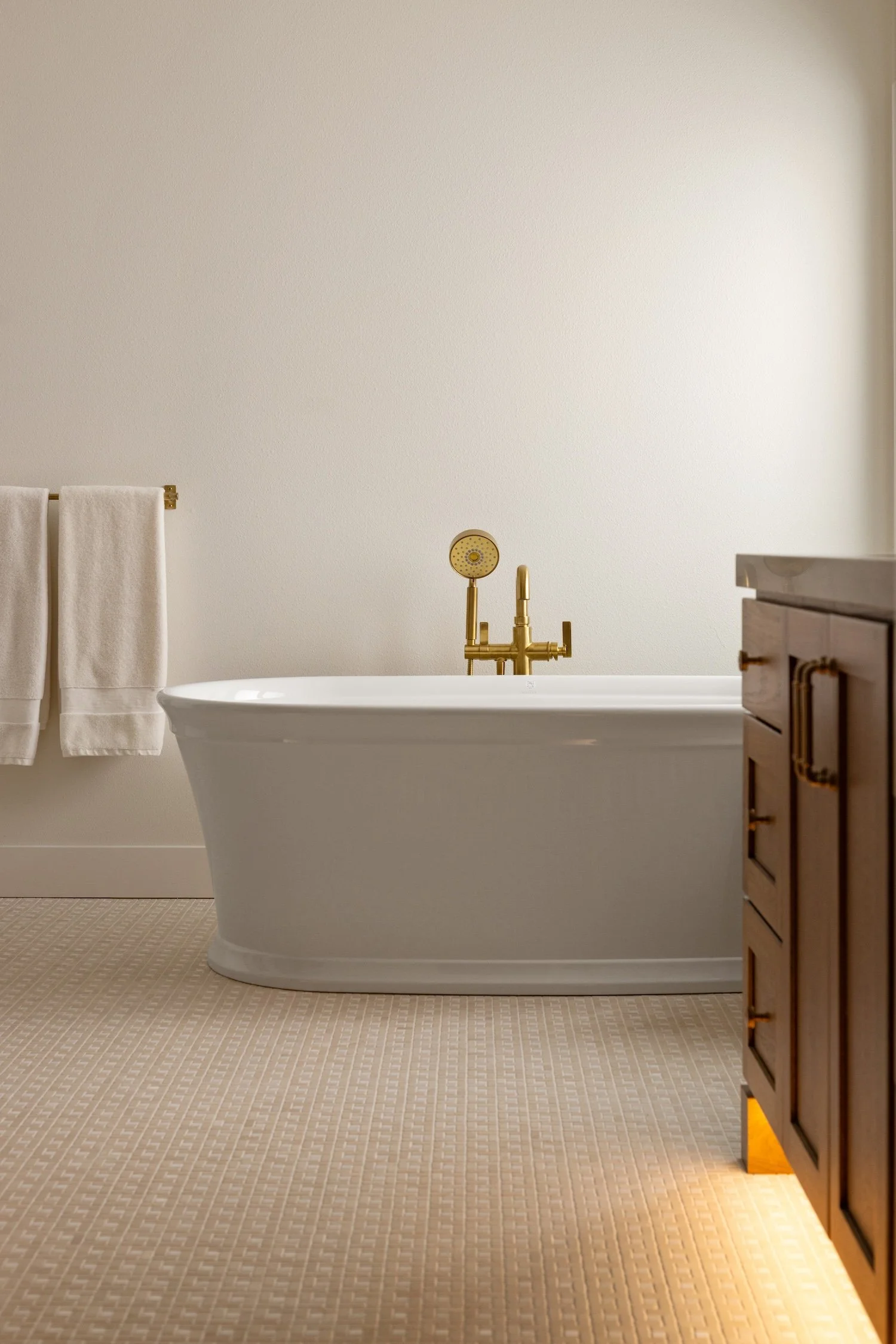 Bathroom with a white bathtub, a golden faucet, and a wooden cabinet on the right. Two white towels hanging on a golden towel rod on the left. The wall is plain and beige with soft lighting.