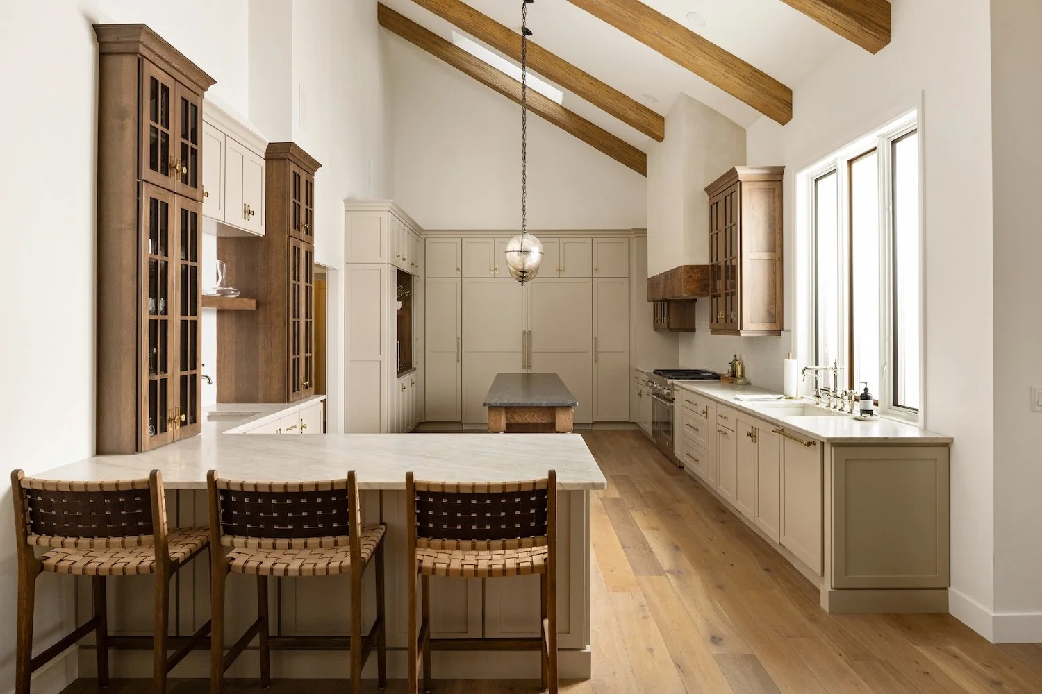 Modern kitchen with white cabinets, wooden accents, and a vaulted ceiling with exposed wooden beams. Features a kitchen island, three woven bar stools, and natural light from large windows.