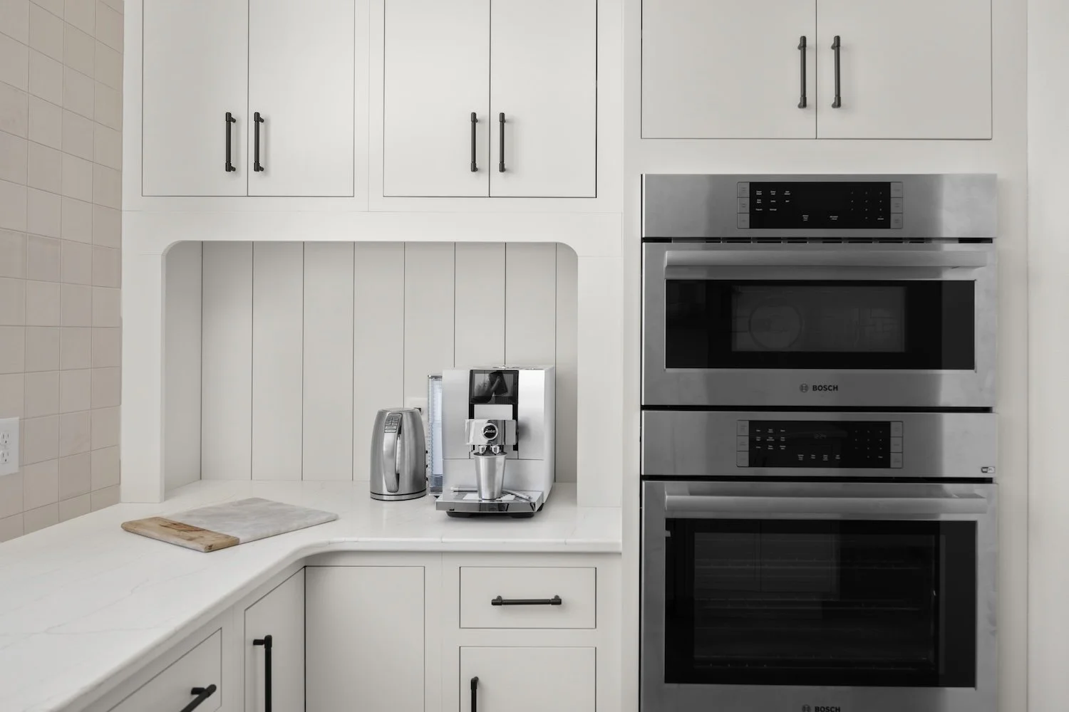 Kitchen counter with coffee maker, electric kettle, and blender; white cabinets, a tile wall, and a stainless steel oven.