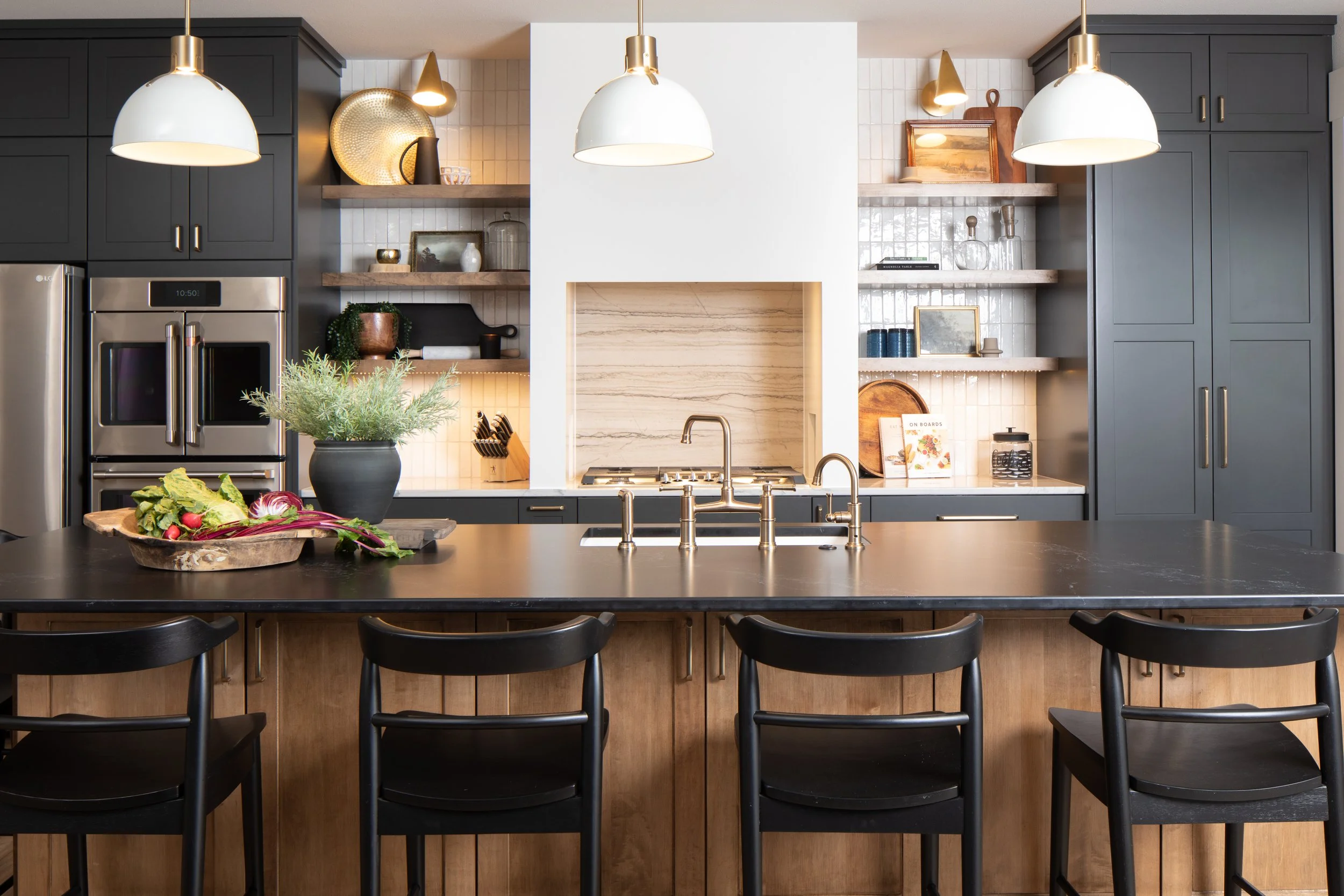 Modern kitchen with dark cabinets, a large island with black chairs, and open shelves with decorative items and kitchenware. Pendant lights hang above the island.