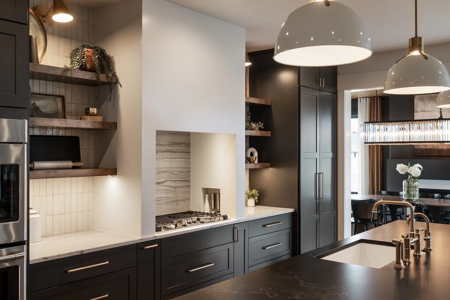 Modern kitchen with dark cabinets, a white marble countertop, and open wooden shelves. Features a built-in gas stove, a stainless steel oven, and hanging pendant lights. In the background, there is a dining area with black chairs and a table with a glass vase of white flowers.