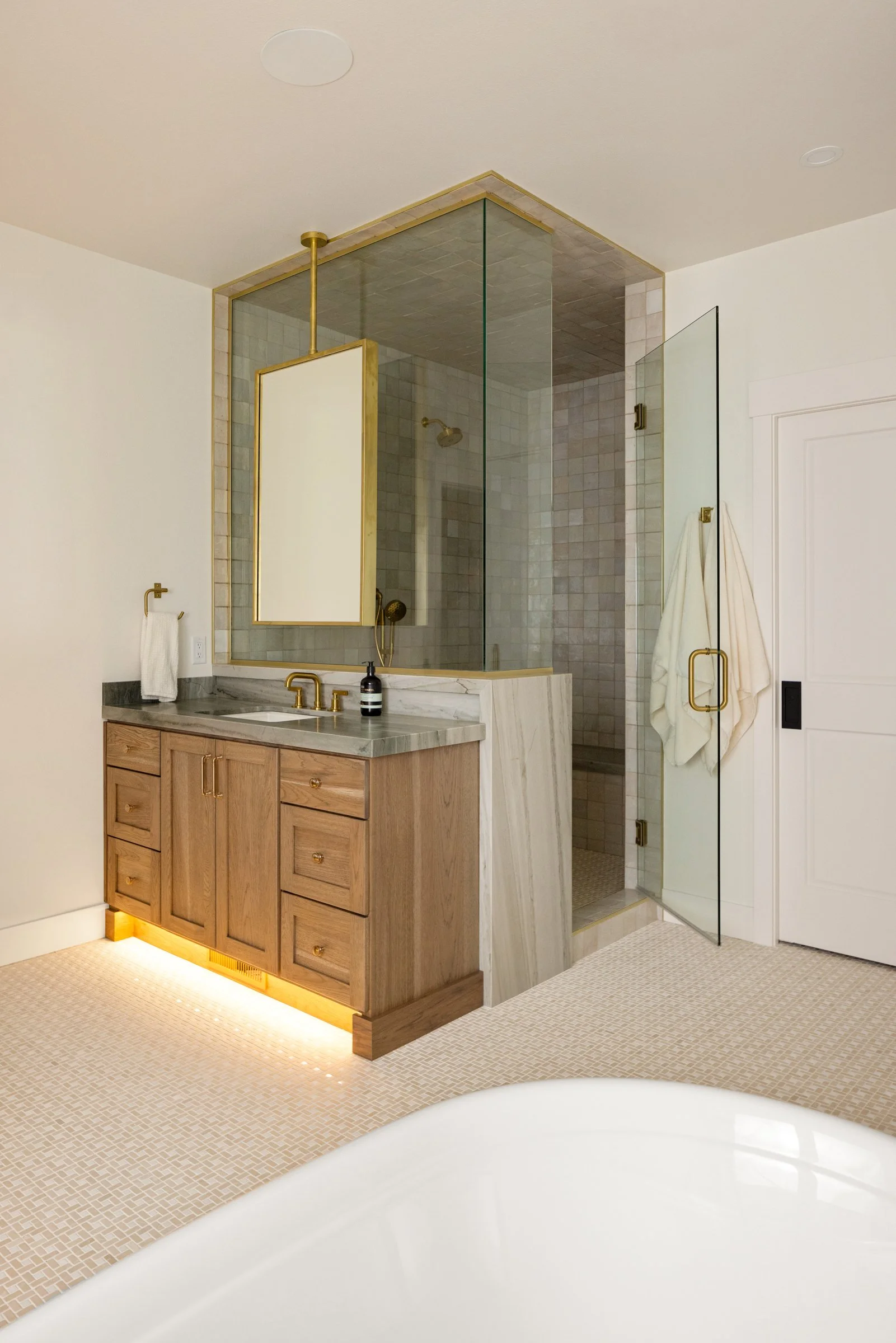 Custom bathroom remodel in Billings, Montana featuring a walk in glass shower with a waterfall stone detail, custom wood vanity, stone countertops, brass fixtures, and refined tilework by Boyce Built.
