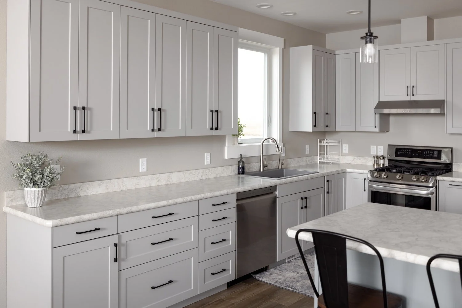 Modern kitchen with white cabinets, stainless steel stove, marble countertops, and a window above the sink, with black cabinet handles and a hanging light fixture.
