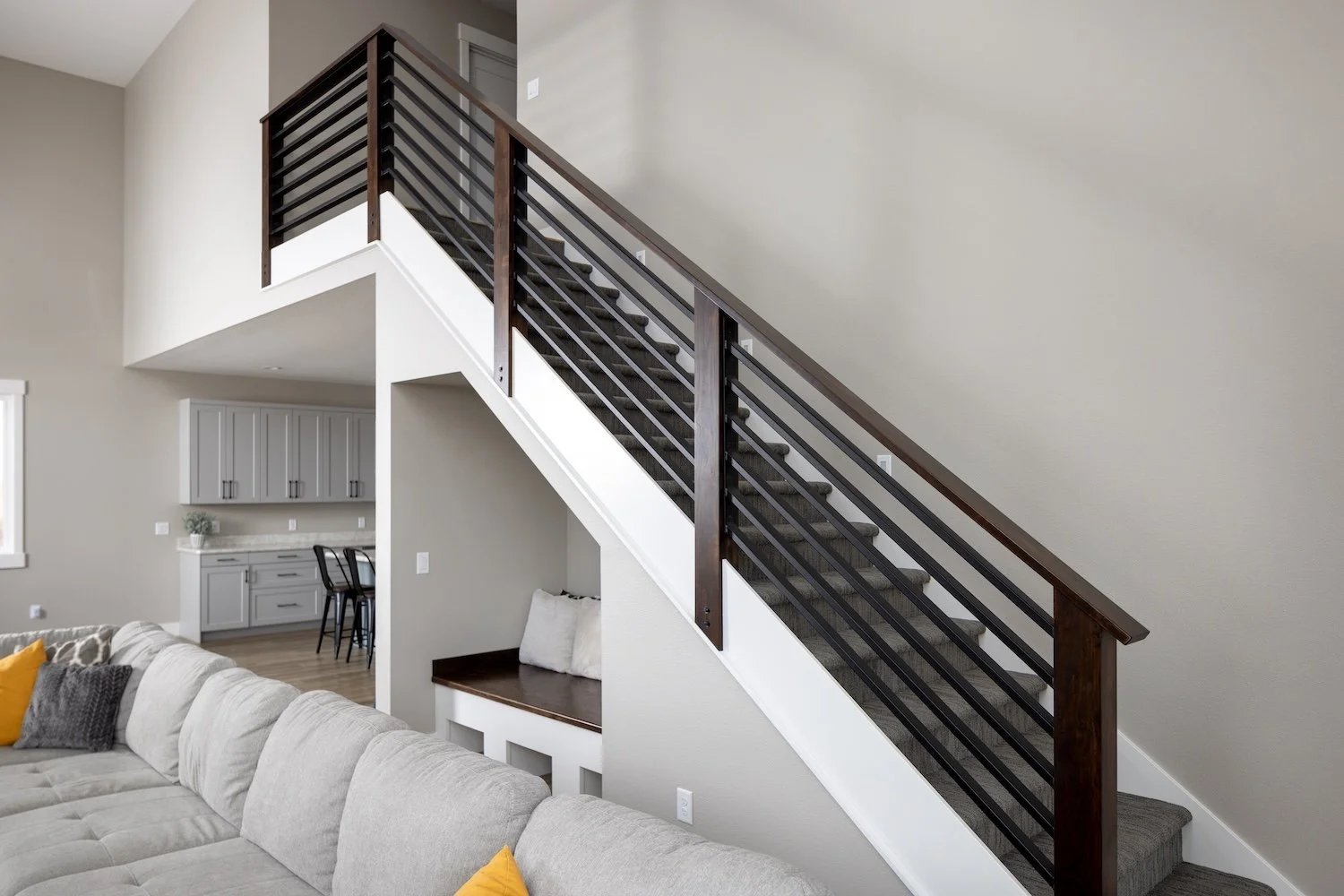 Interior of a modern living room with beige walls and a staircase with dark wood and black metal railings. Part of a light-colored couch with pillows, and a built-in nook under the staircase with cushions and a small seating area visible.