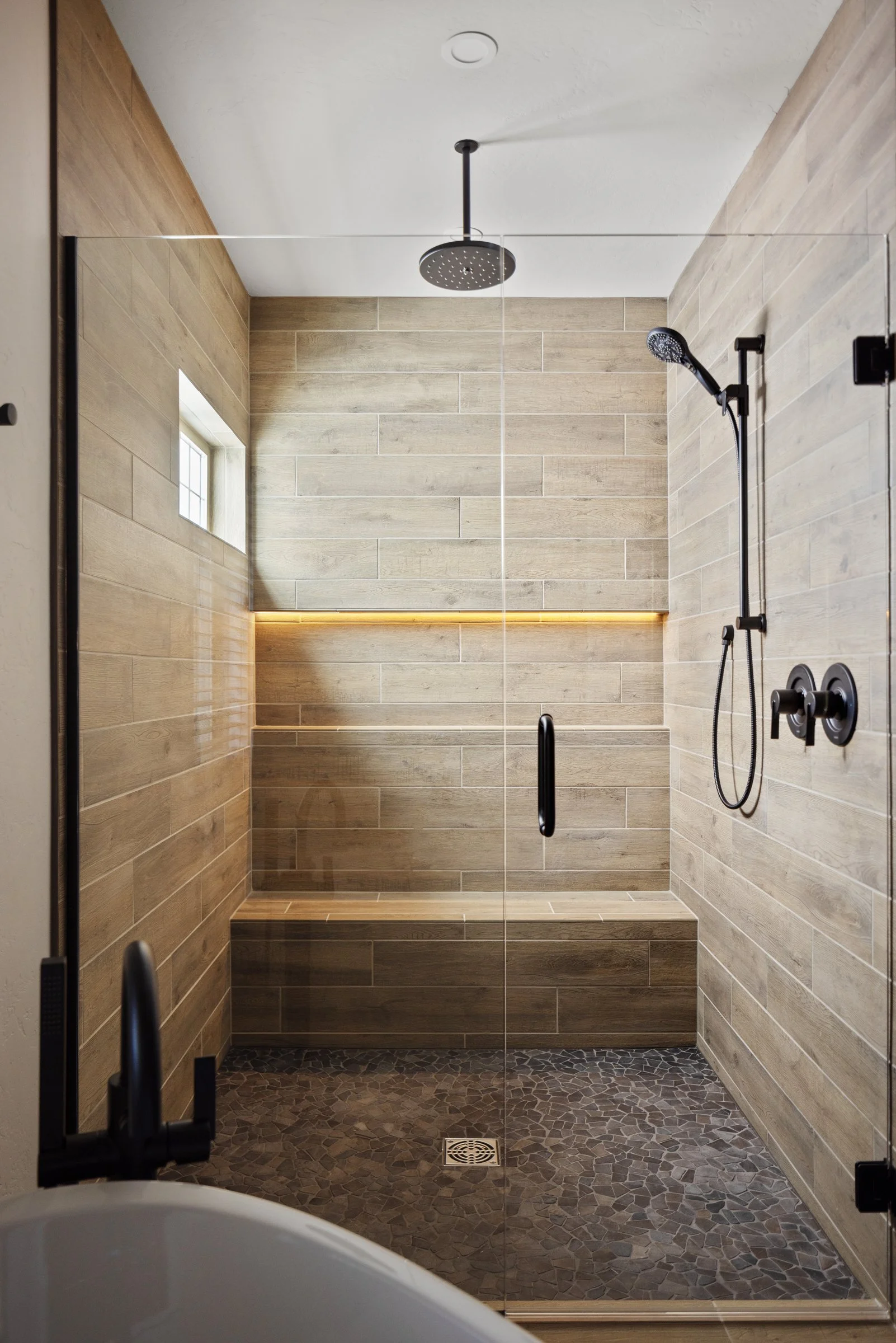 Modern walk-in shower with wood-look tiled walls and pebble stone floor, featuring a rain showerhead, handheld shower, built-in bench, small window, and a glass door.