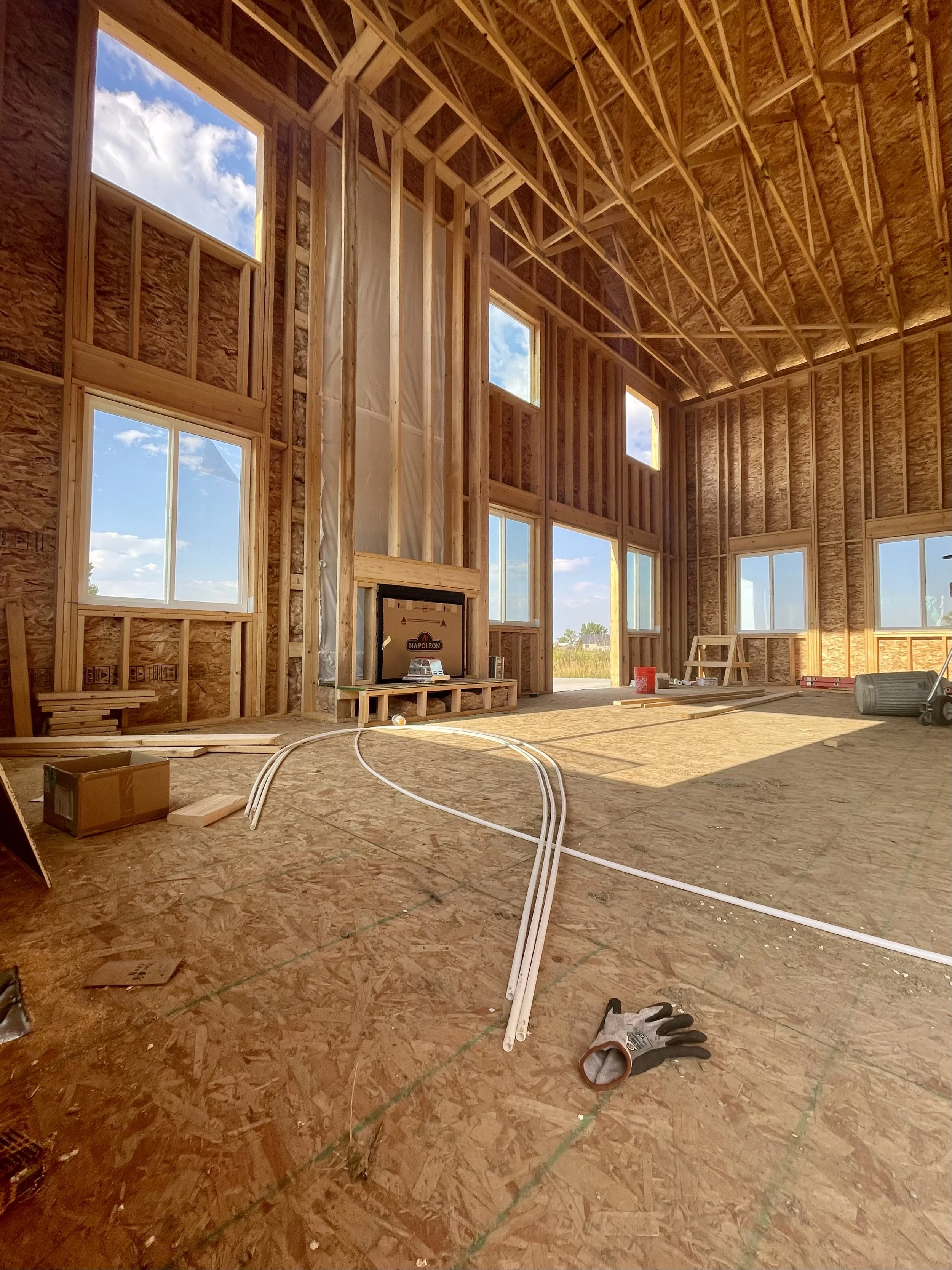 Interior of a house under construction with exposed wooden framing, several windows, construction tools, and materials on the floor.