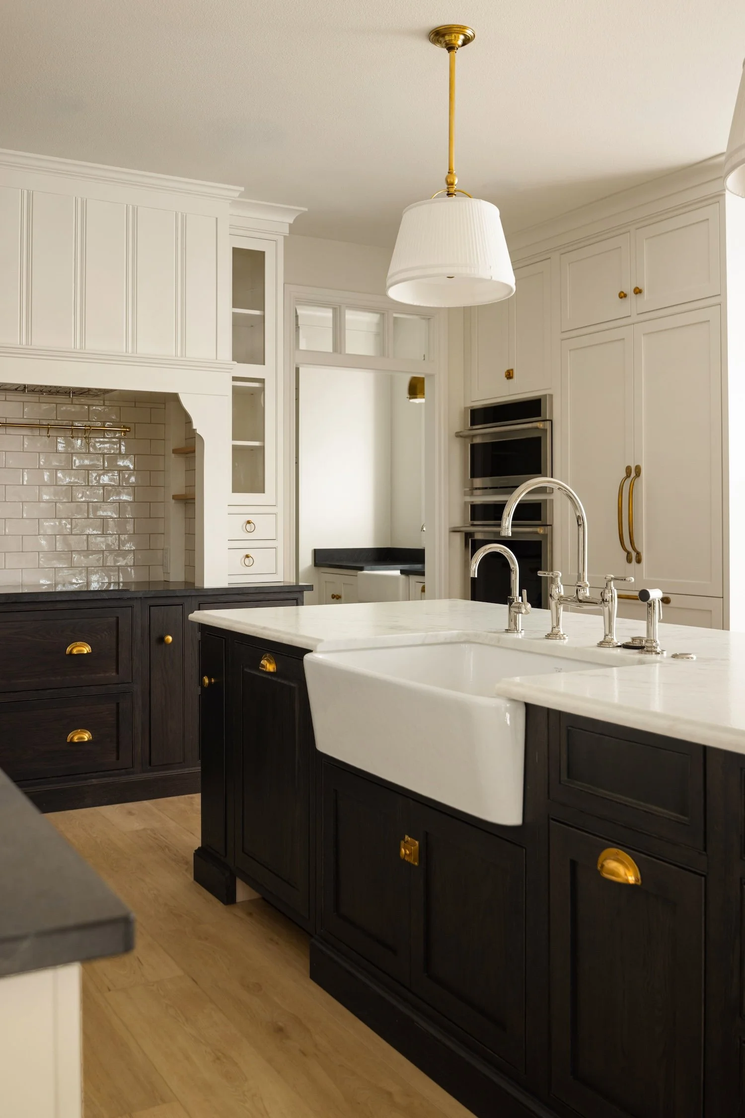 A modern kitchen with a white island sink, black cabinetry with gold hardware, a white marble countertop, white cabinets with gold knobs, and a white tiled backsplash. Overhead is a white pendant light with gold accents. There are wooden floors.