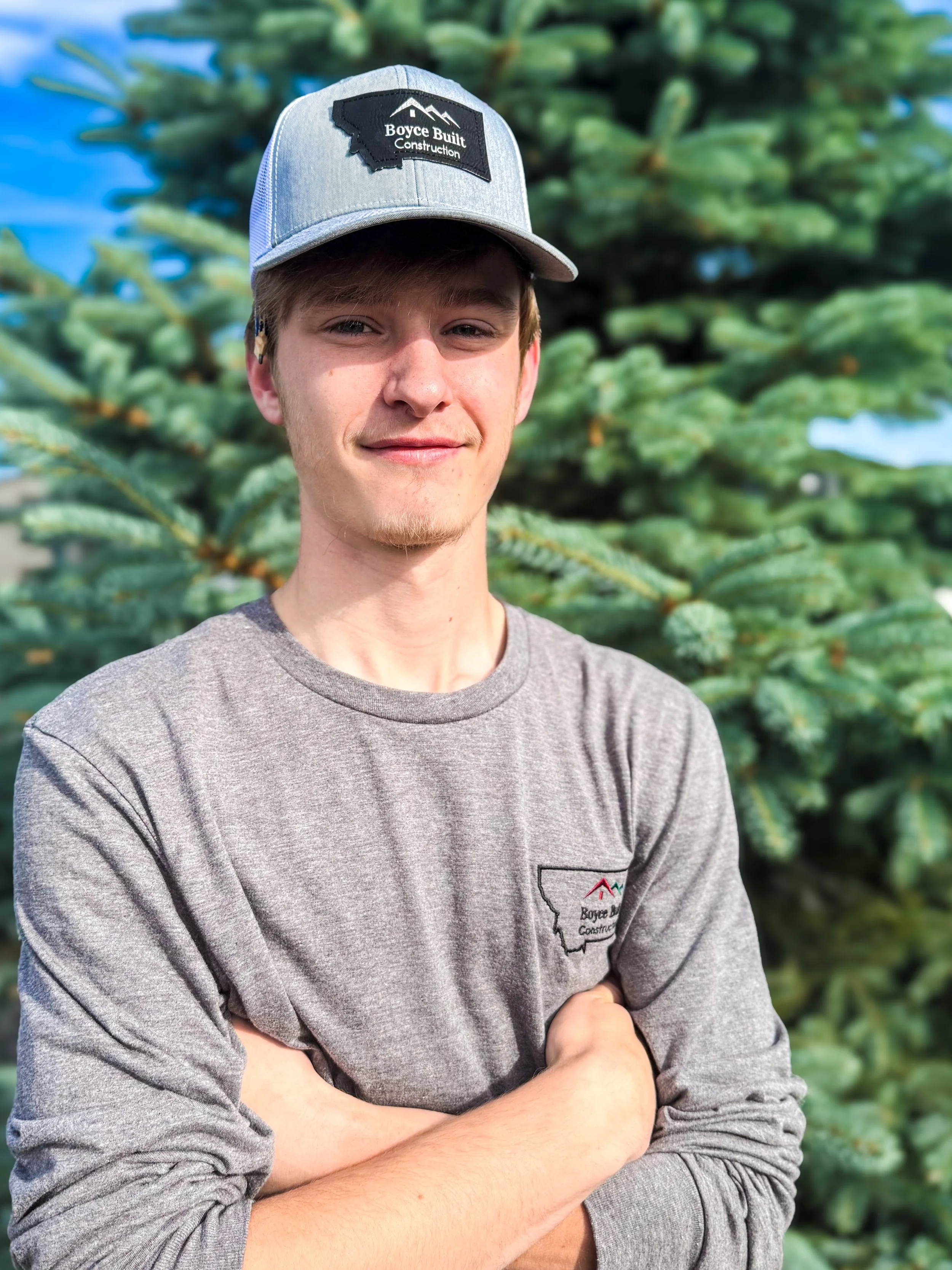 Young man wearing a gray cap and shirt branded with Boyce Built Construction, standing in front of a pine tree with arms crossed.