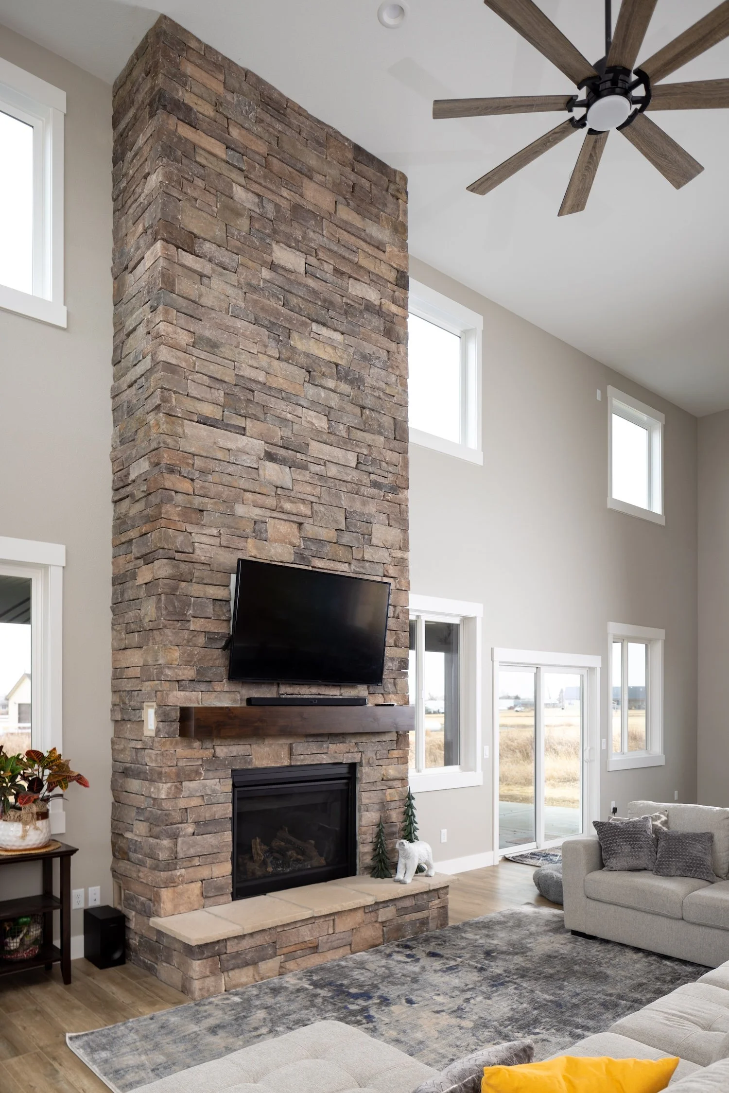 Living room with a brick fireplace, mounted TV, gray sofa with yellow pillows, wood floor, large windows, sliding door, ceiling fan, and area rug.