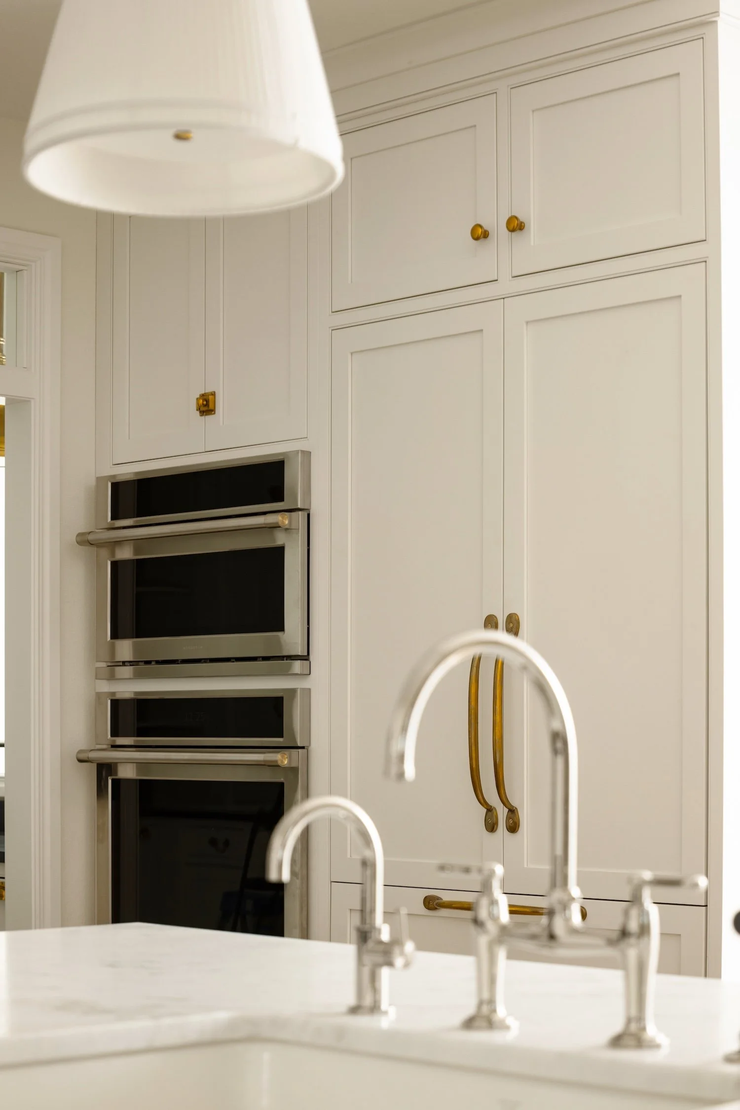 White kitchen with built-in oven, white cabinets with gold knobs, gold hardware, and a white countertop with a modern faucet.