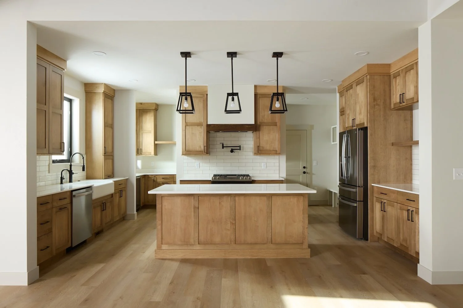Modern kitchen with wooden cabinets, white countertops, black pendant lights, white backsplash, stainless steel appliances, and hardwood flooring.
