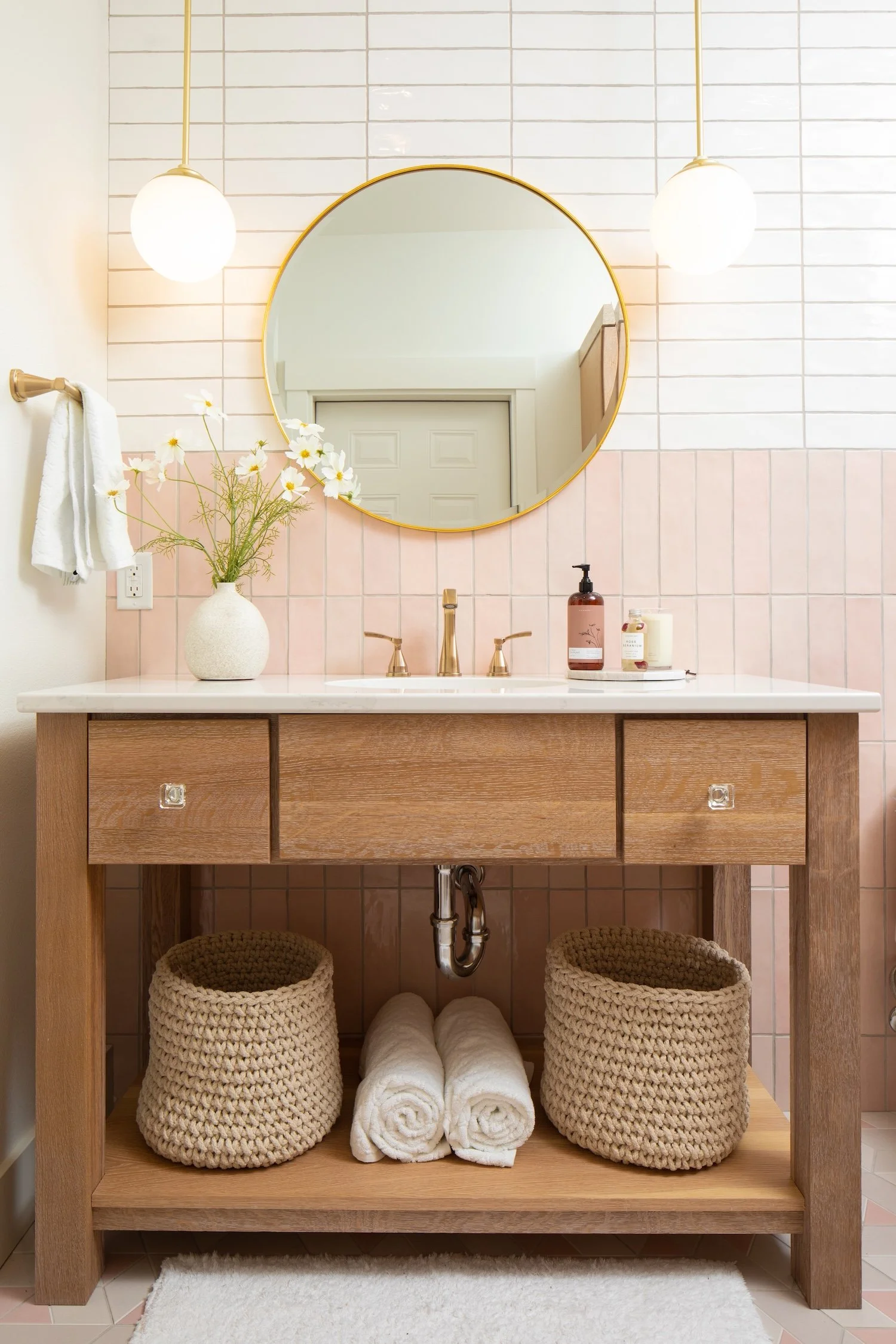 Bathroom vanity with a marble countertop, gold faucet, and a mirror in a round gold frame. Decor includes a white vase with white flowers, soap dispenser, candle, and rolled towels. Two woven baskets and rolled towels are stored underneath on a wooden shelf. The wall is tiled with light pink tiles.