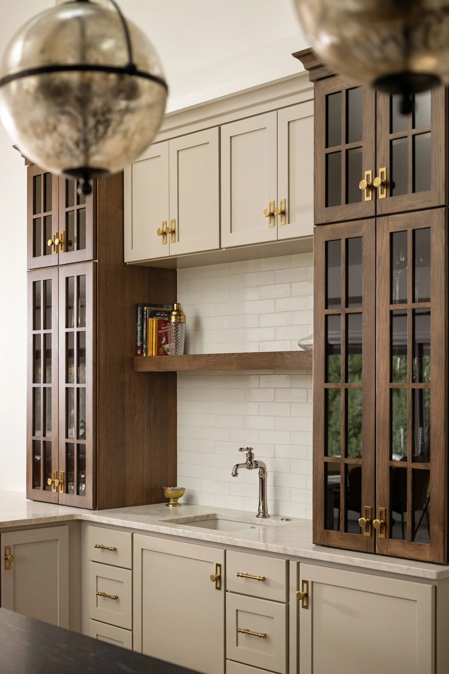 Kitchen with white lower cabinets, beige upper cabinets, dark wooden display cabinets with glass doors, and gold hardware. There is a small shelf with books and a gold-colored bottle, a white subway tile backsplash, and a silver faucet over a white sink.