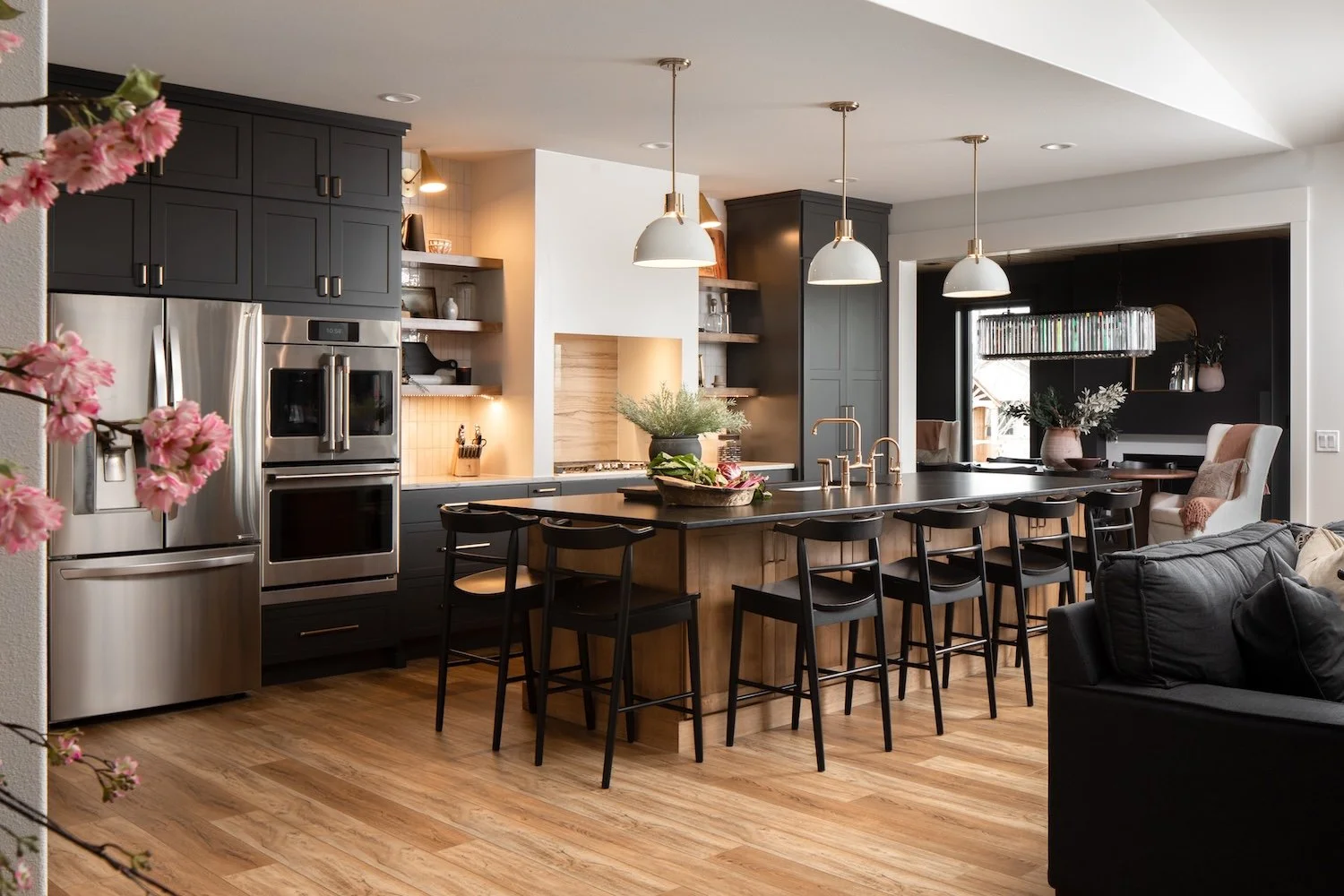 Modern kitchen with dark cabinetry, stainless steel appliances, a large island with seating, and open shelving decorated with plants and dishes, adjacent to a cozy living area.