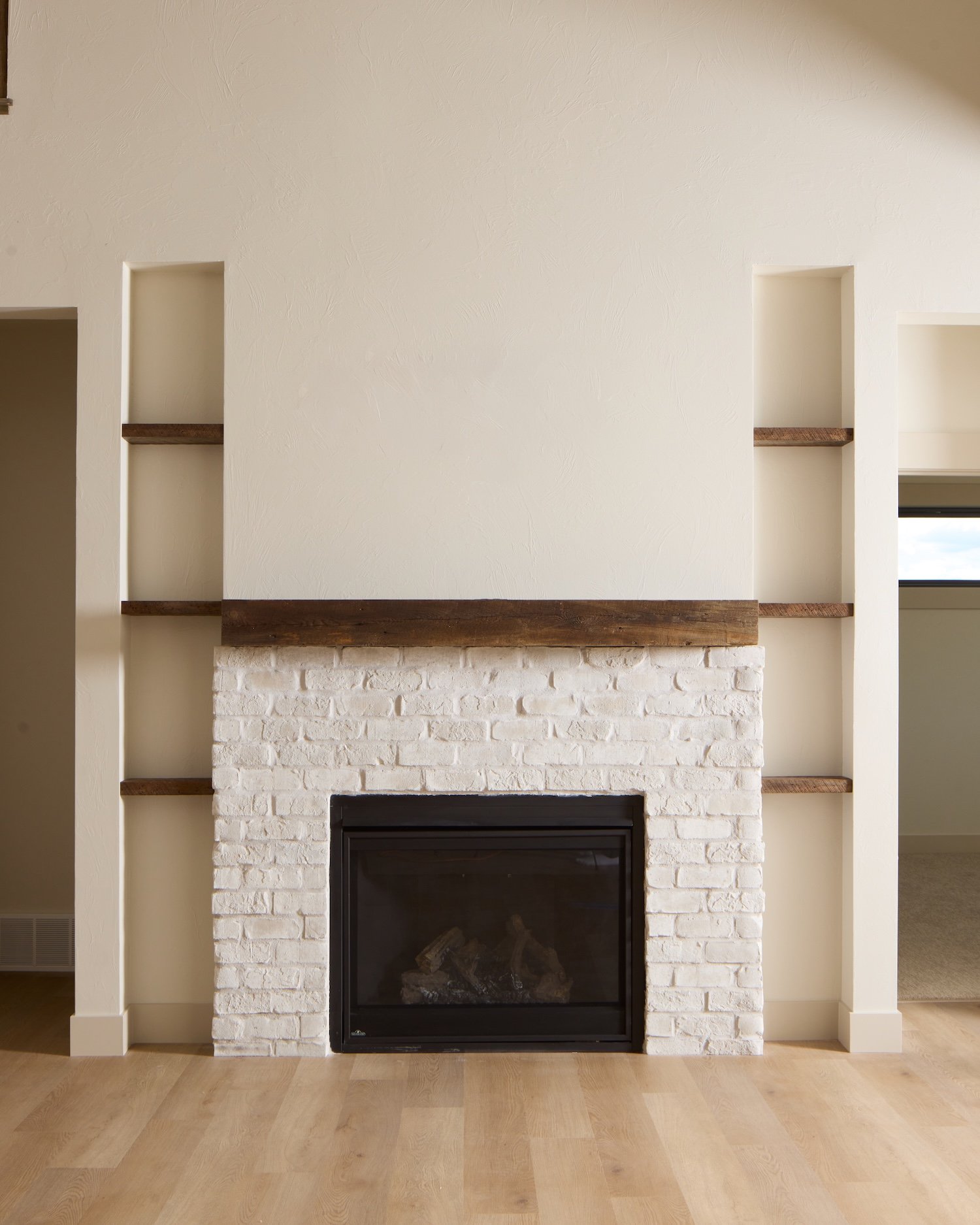 A white brick fireplace with a black metal frame, surrounded by built-in wall shelves with wooden shelves on either side. The wall above the fireplace is painted in a light beige color.