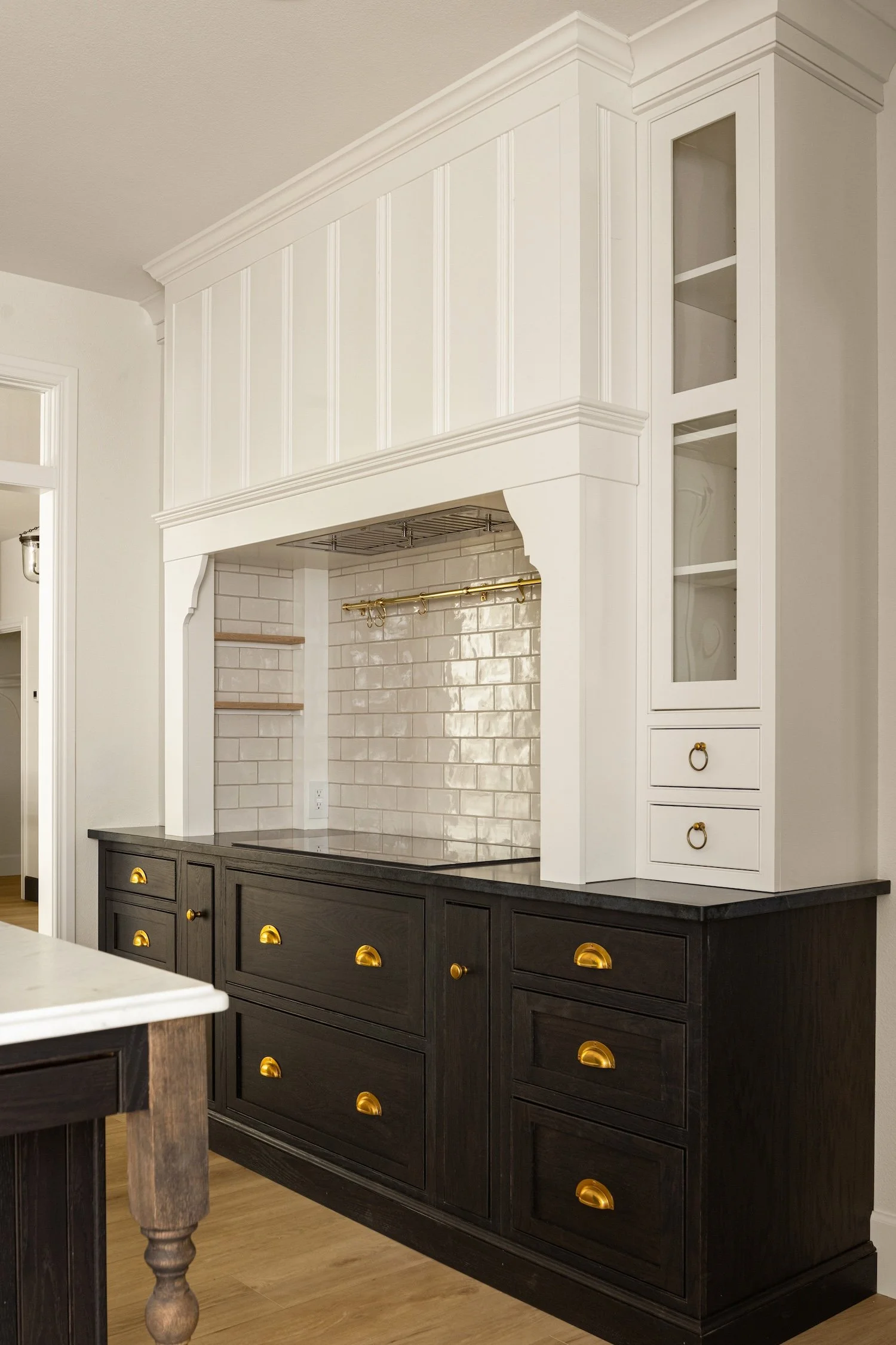 A kitchen with a black lower cabinet and a white upper cabinet with glass doors, gold drawer pulls and cabinet handles, a white tile backsplash, and a white countertop. The lower cabinet has multiple drawers.