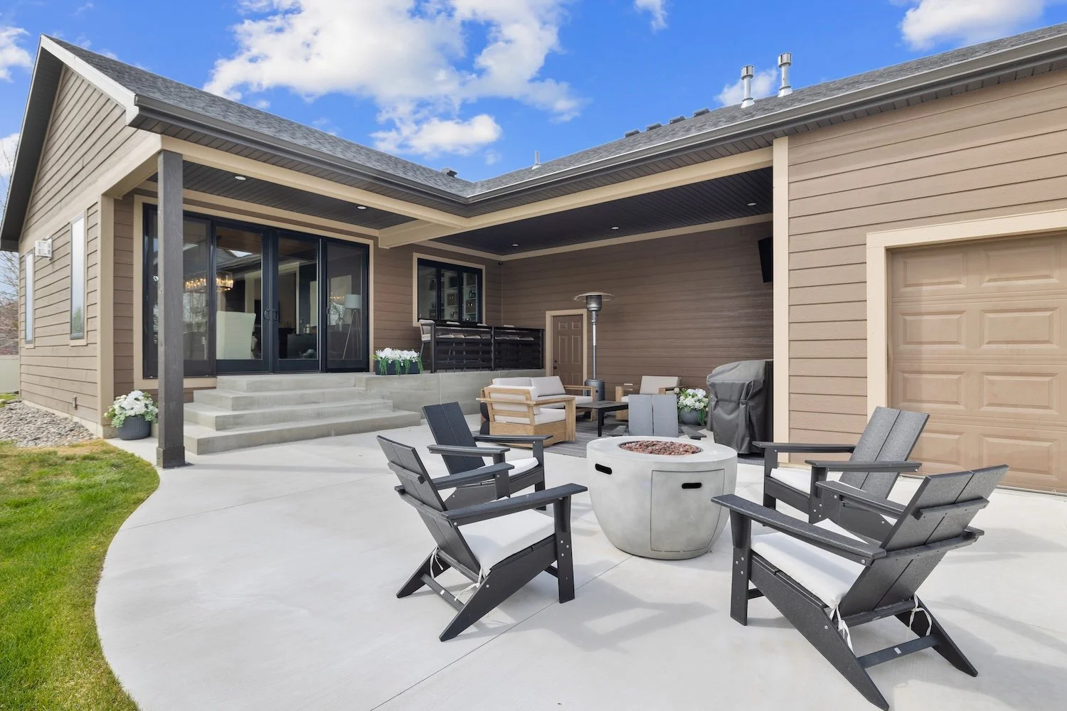 A modern outdoor patio with chairs, a fire pit, and a covered porch area attached to a house with siding, windows, and a garage door.