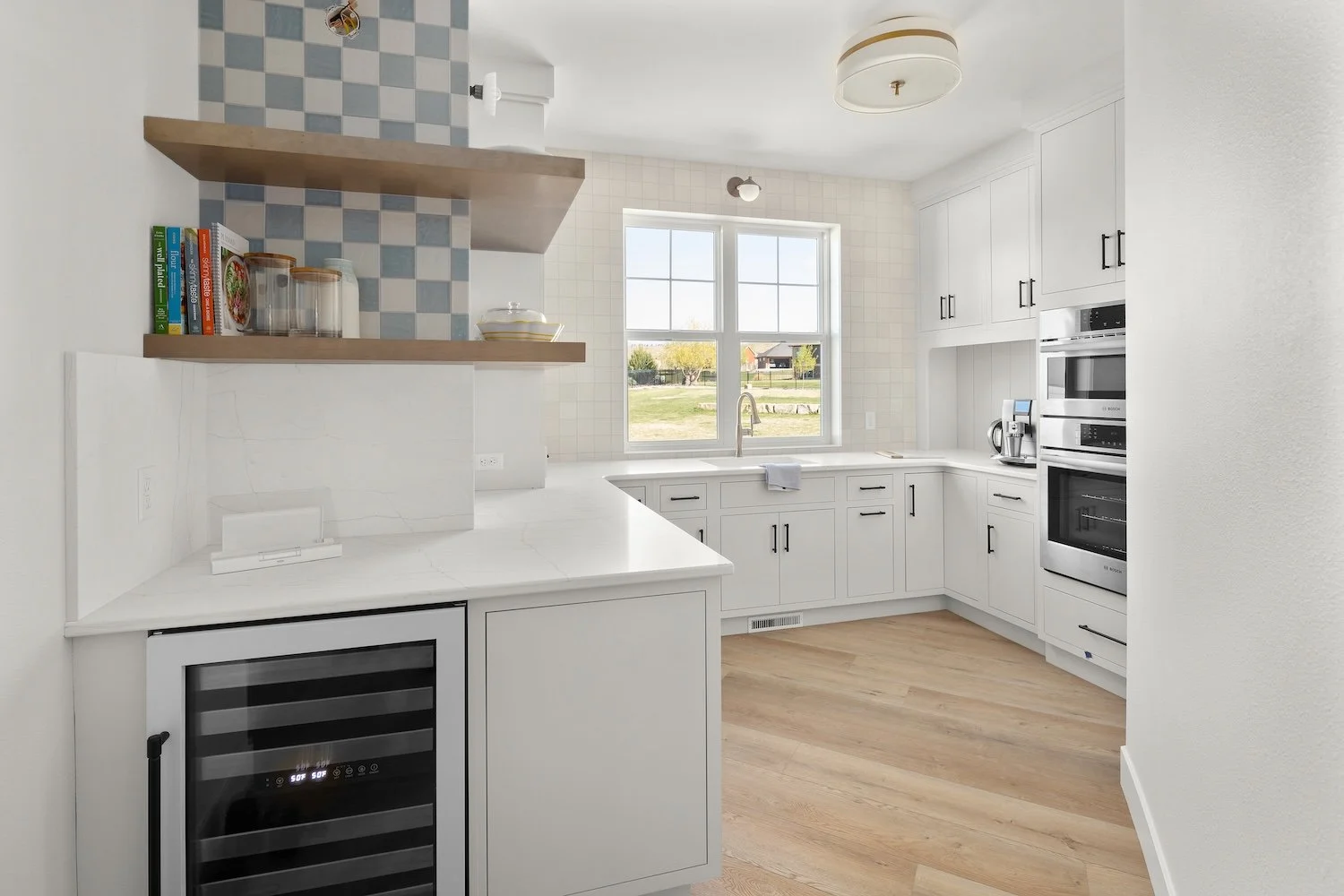 Bright kitchen with white cabinets, a window overlooking a yard, and built-in appliances including an oven and coffee maker. Wooden flooring and a backsplash with blue and white checkered tiles.