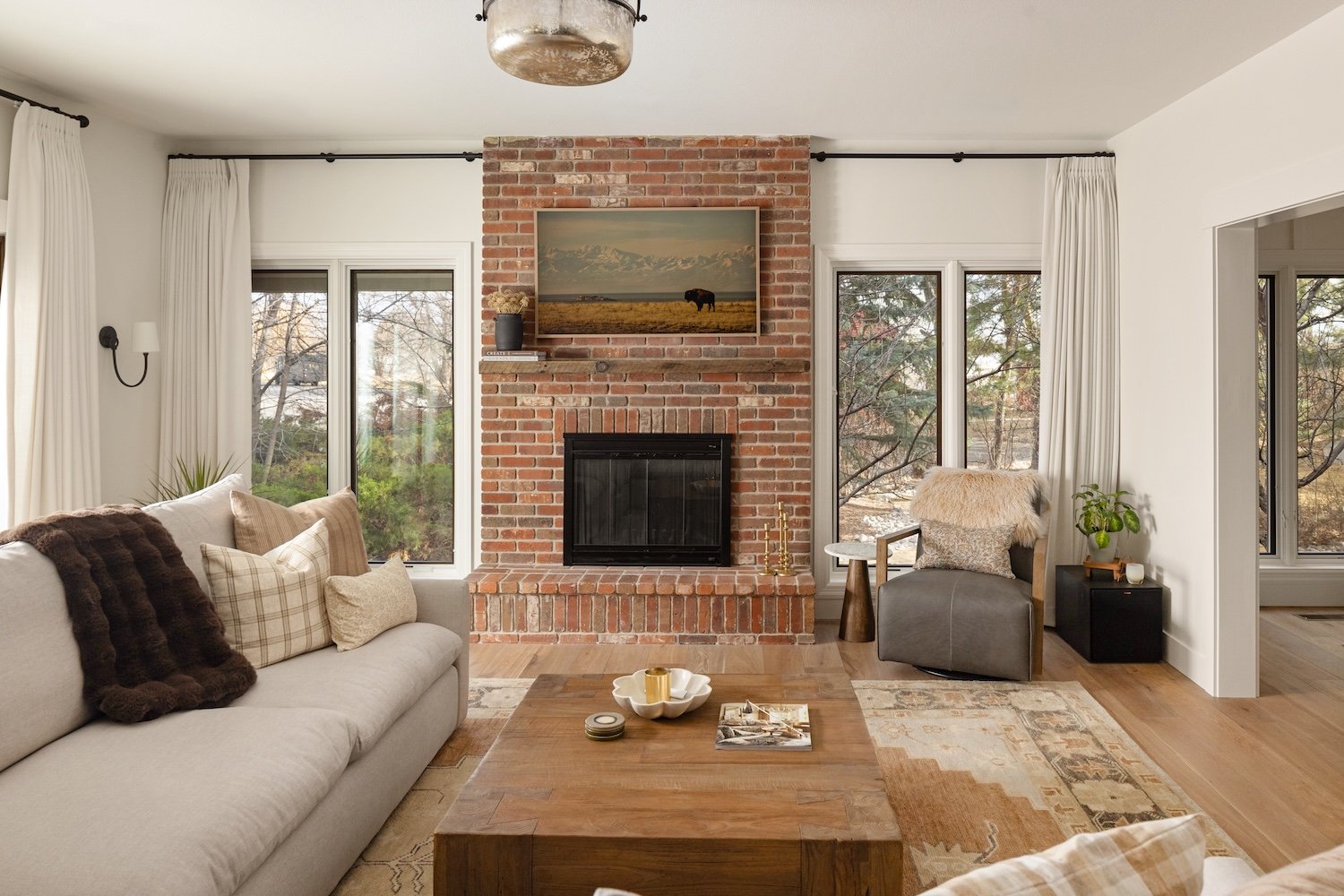 Living room with a brick fireplace, a beige sofa with plaid and furry pillows, a wooden coffee table with candles, and a gray armchair with a fur cushion, surrounded by windows and white curtains.