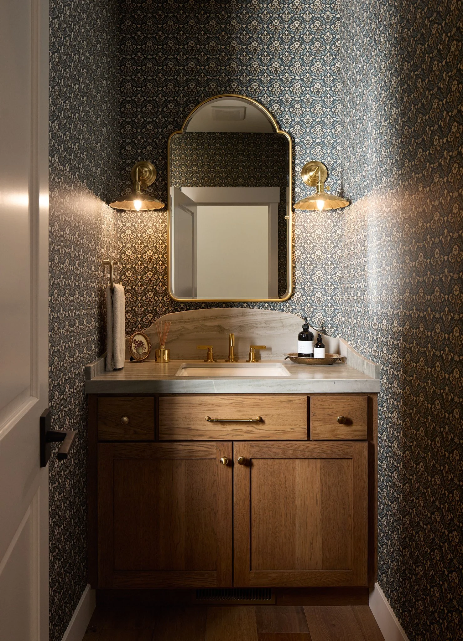 Custom powder room remodel in Billings, Montana featuring warm wood cabinetry, marble countertops, brass fixtures, patterned wallpaper, and intentional custom detailing by Boyce Built.