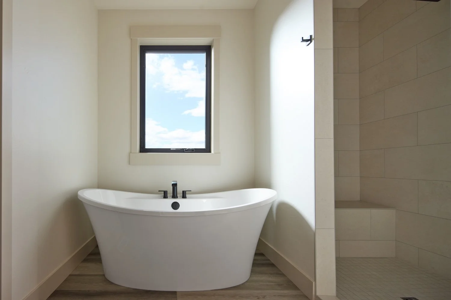 Modern bathroom featuring a white freestanding bathtub under a window with a view of the blue sky and clouds, next to a tiled shower area.
