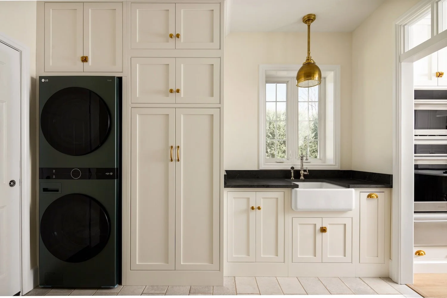 Kitchen with a stacked LG washer and dryer, white cabinets with brass handles, a black countertop, a white farmhouse sink, a window above the sink, a gold pendant light, and a pantry on the right.