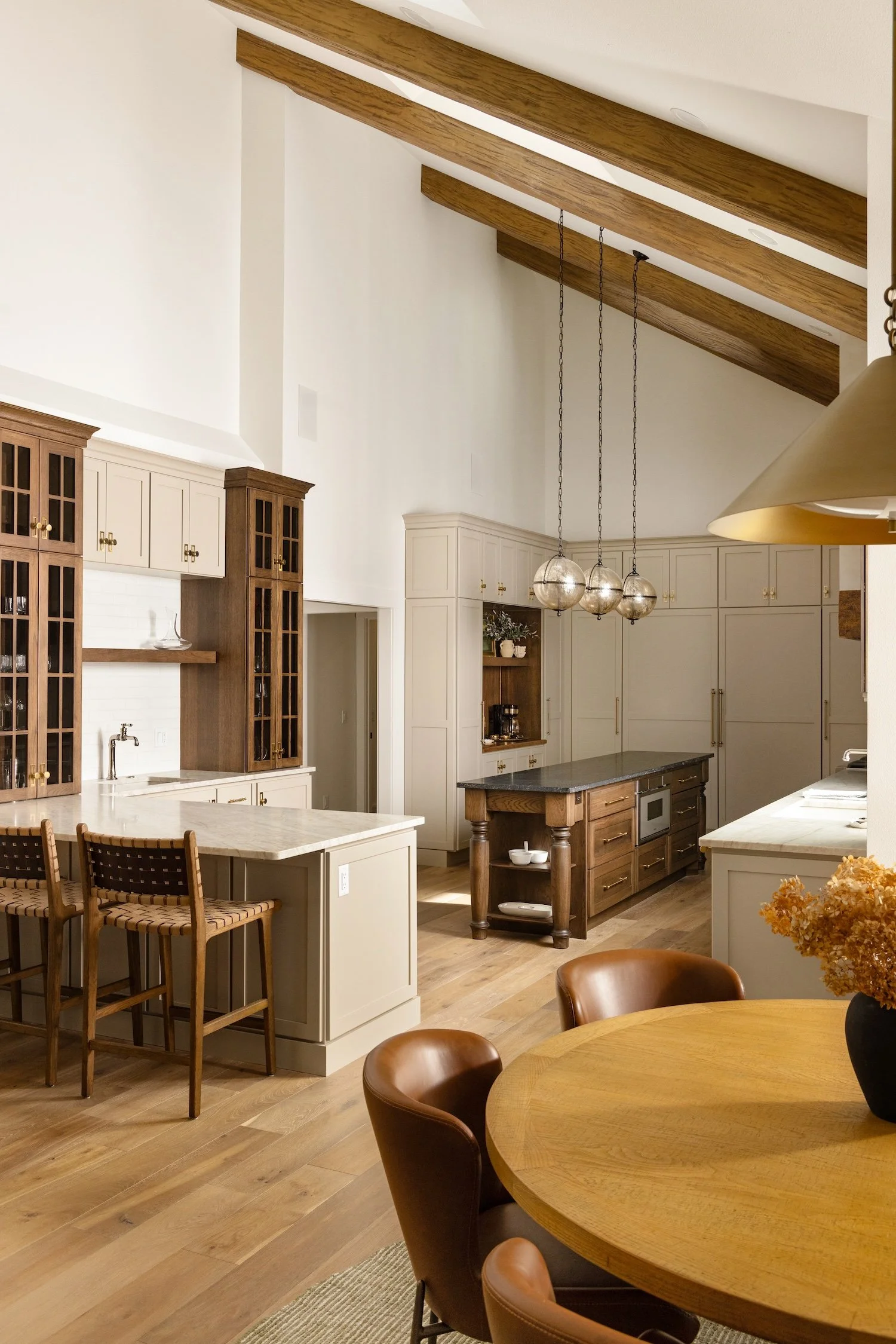A modern kitchen with white and wooden cabinets, a kitchen island, and a dining area with a round wooden table and brown leather chairs. There are ceiling beams and hanging spherical pendant lights.