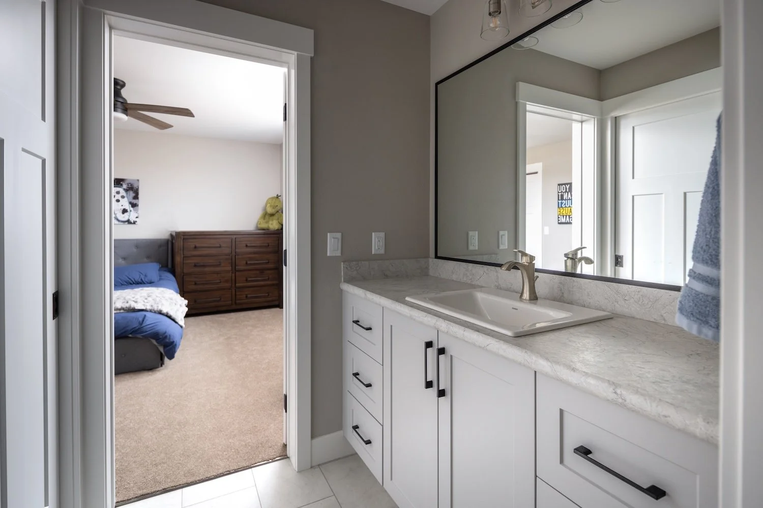 Bathroom sink with a large mirror, marble countertop, and a view into a bedroom with a bed, dresser, and stuffed animal.