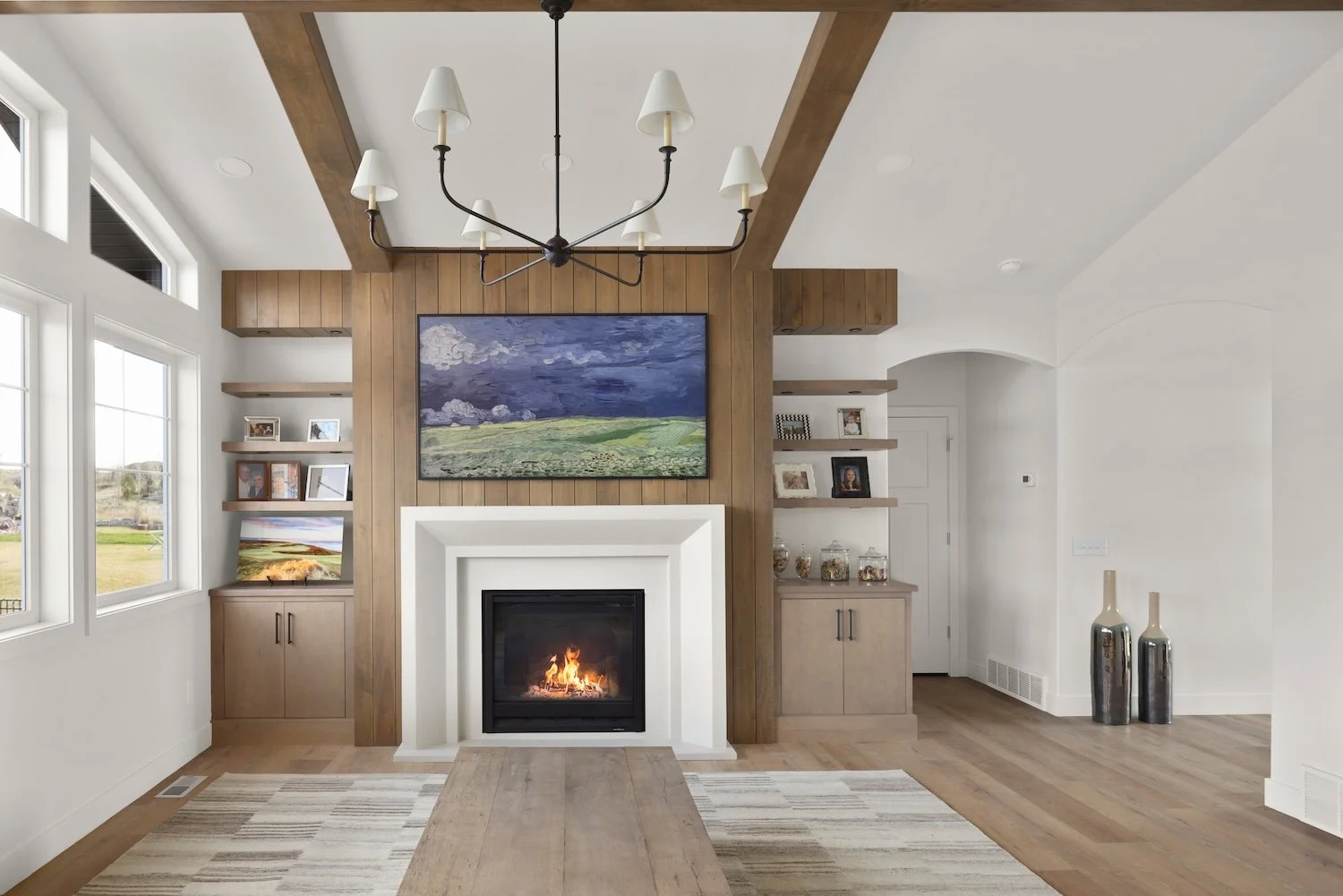 Living room with a fireplace, wooden accents, large windows, a chandelier, and artwork. Decorative vases are on the floor.